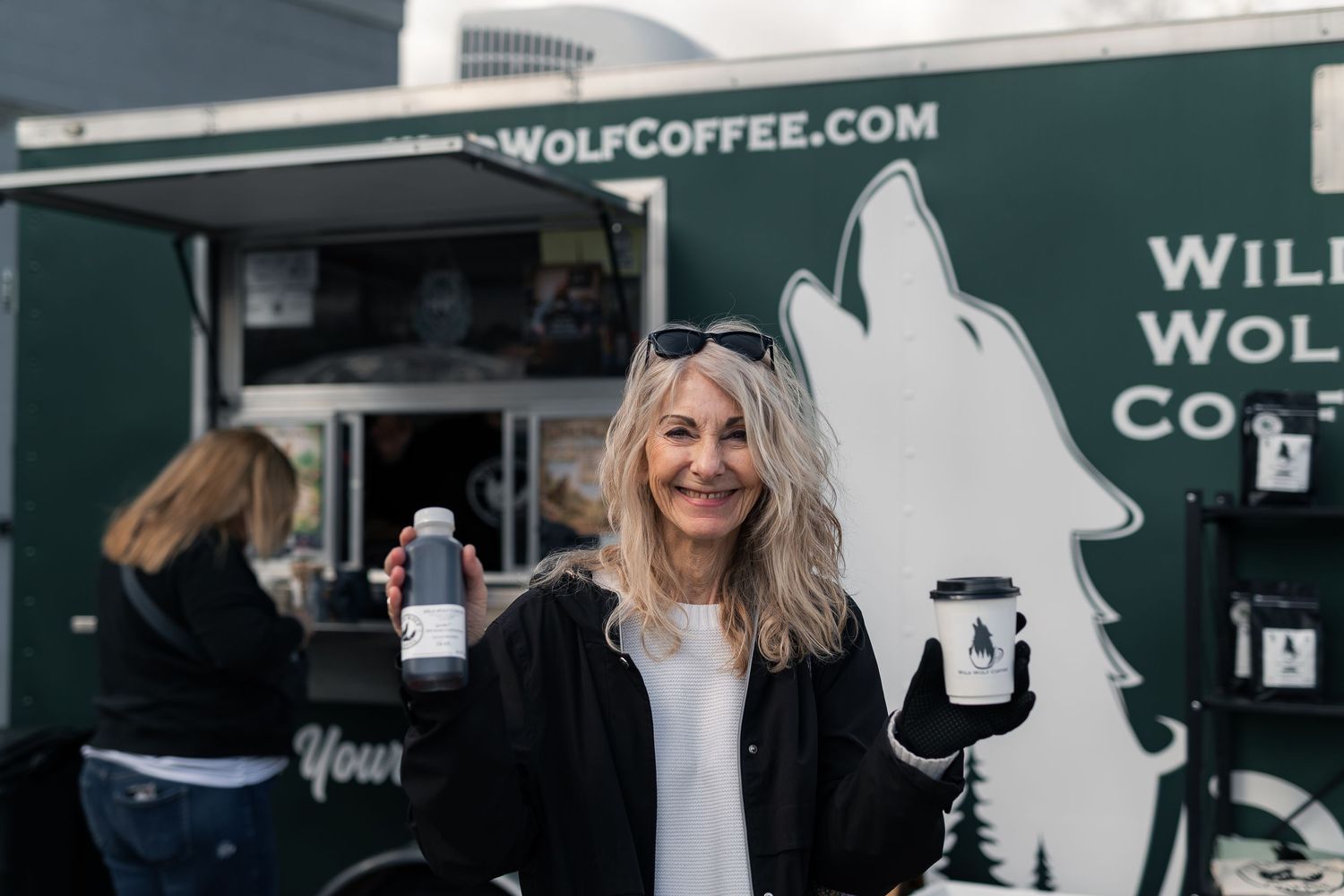Smiling woman holding Wild Wolf Coffee cups outside a dark green Wild Wolf Coffee food truck.