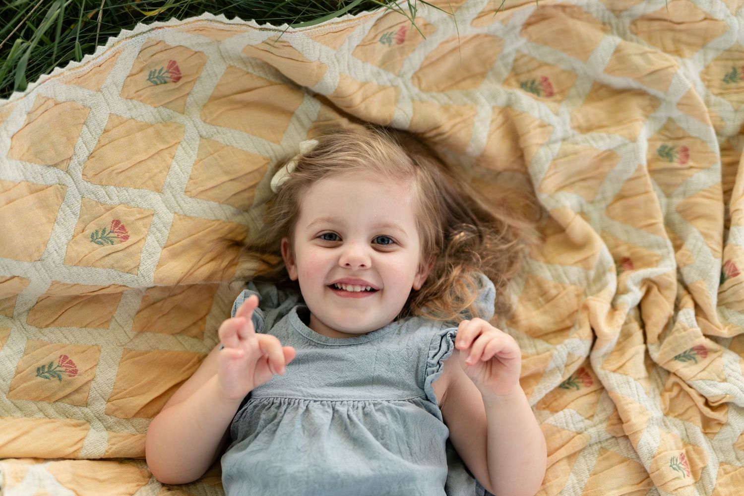 Smiling toddler girl in gray dress lying on yellow floral quilt outdoors.