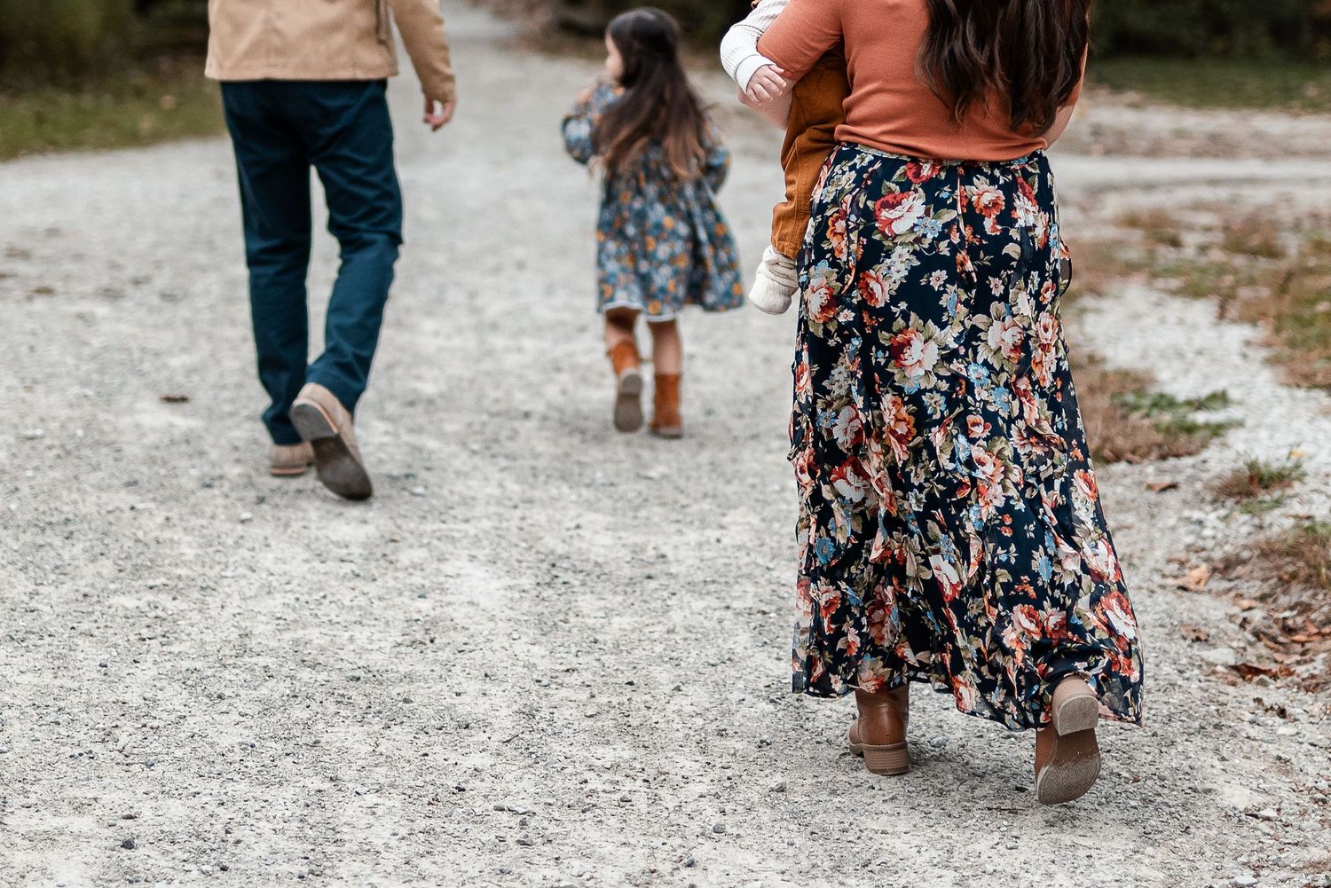 Family walking away on a gravel path, with a woman in a floral skirt carrying a child and a man in jeans beside a little girl in boots.