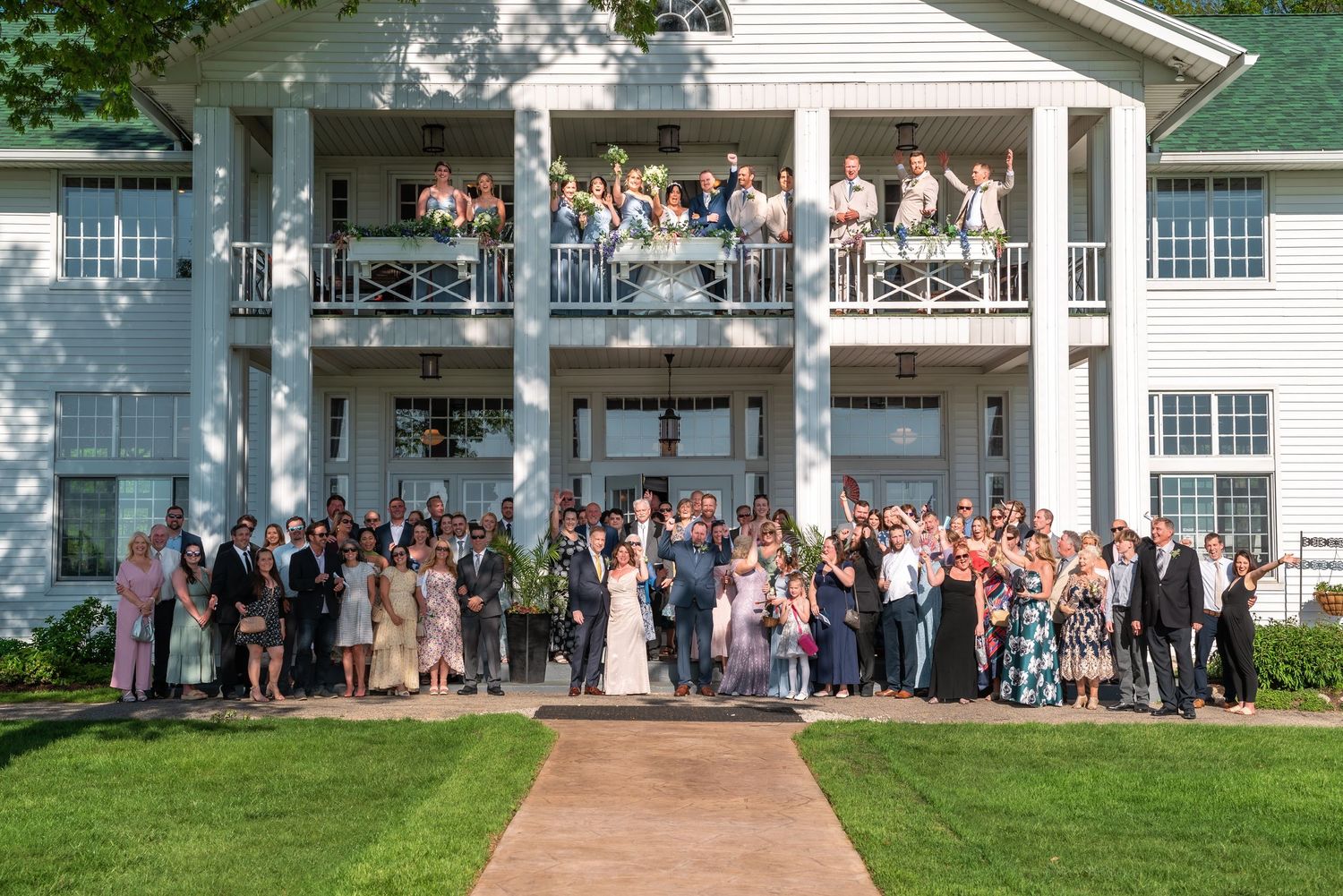 Large wedding party gathered on the lawn in front of a white two-story colonial mansion with wrap-around porch.