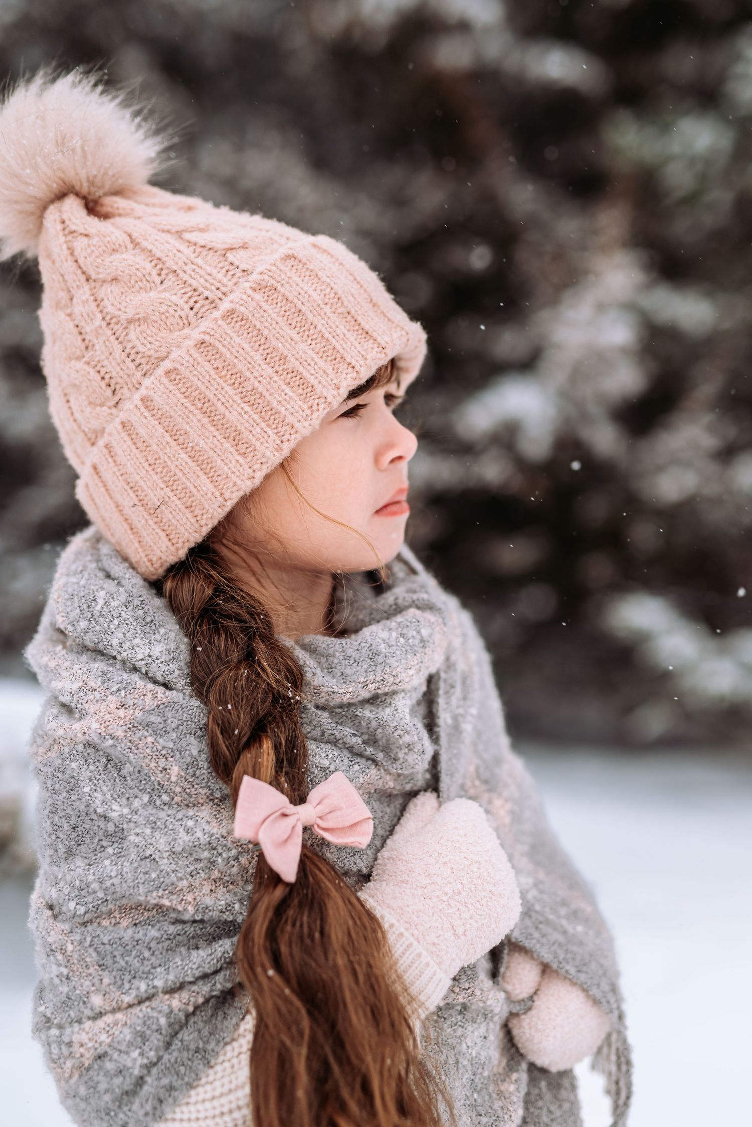 A cozy winter ensemble with a pink bow detail, knit hat, and gray scarf against a snowy background.