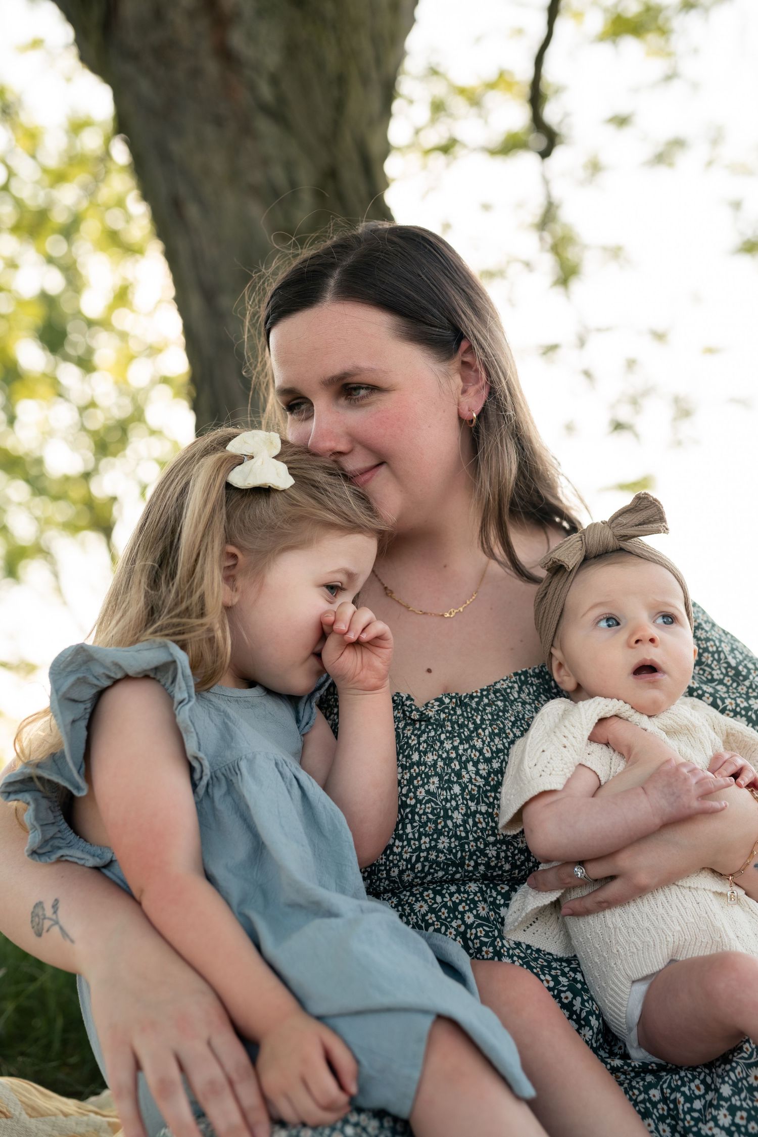 A mother sits outdoors under a tree, lovingly holding her toddler girl and baby in a warm family portrait.