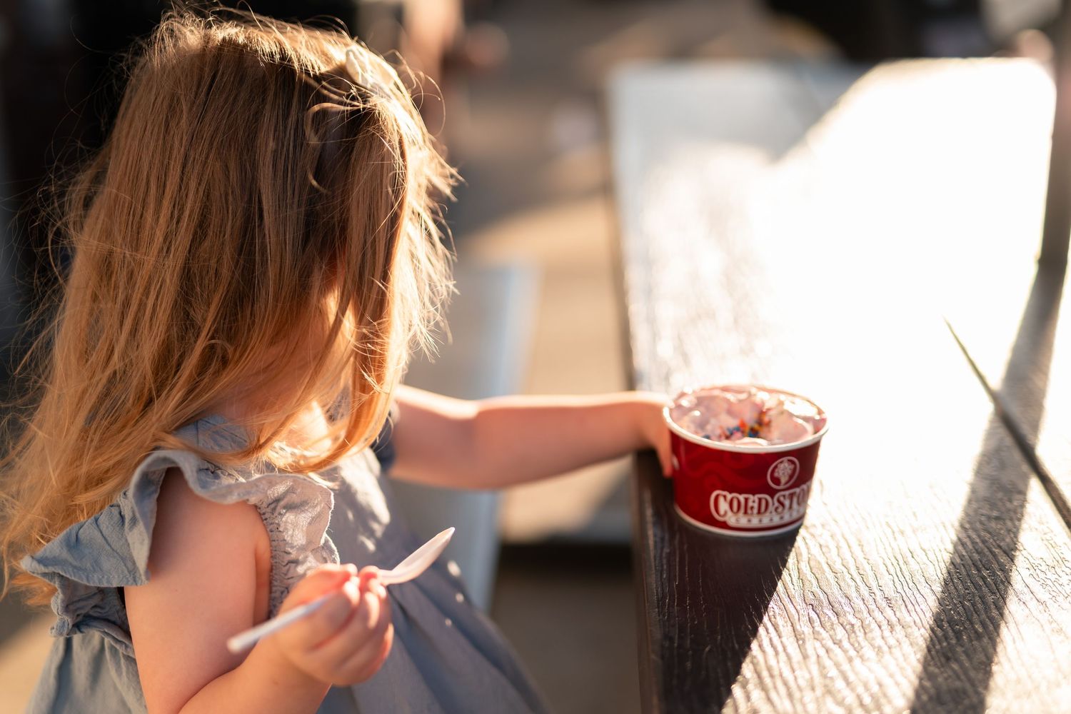 Young girl in gray dress reaching for a red Cold Stone Creamery drink cup on a sunny outdoor table.