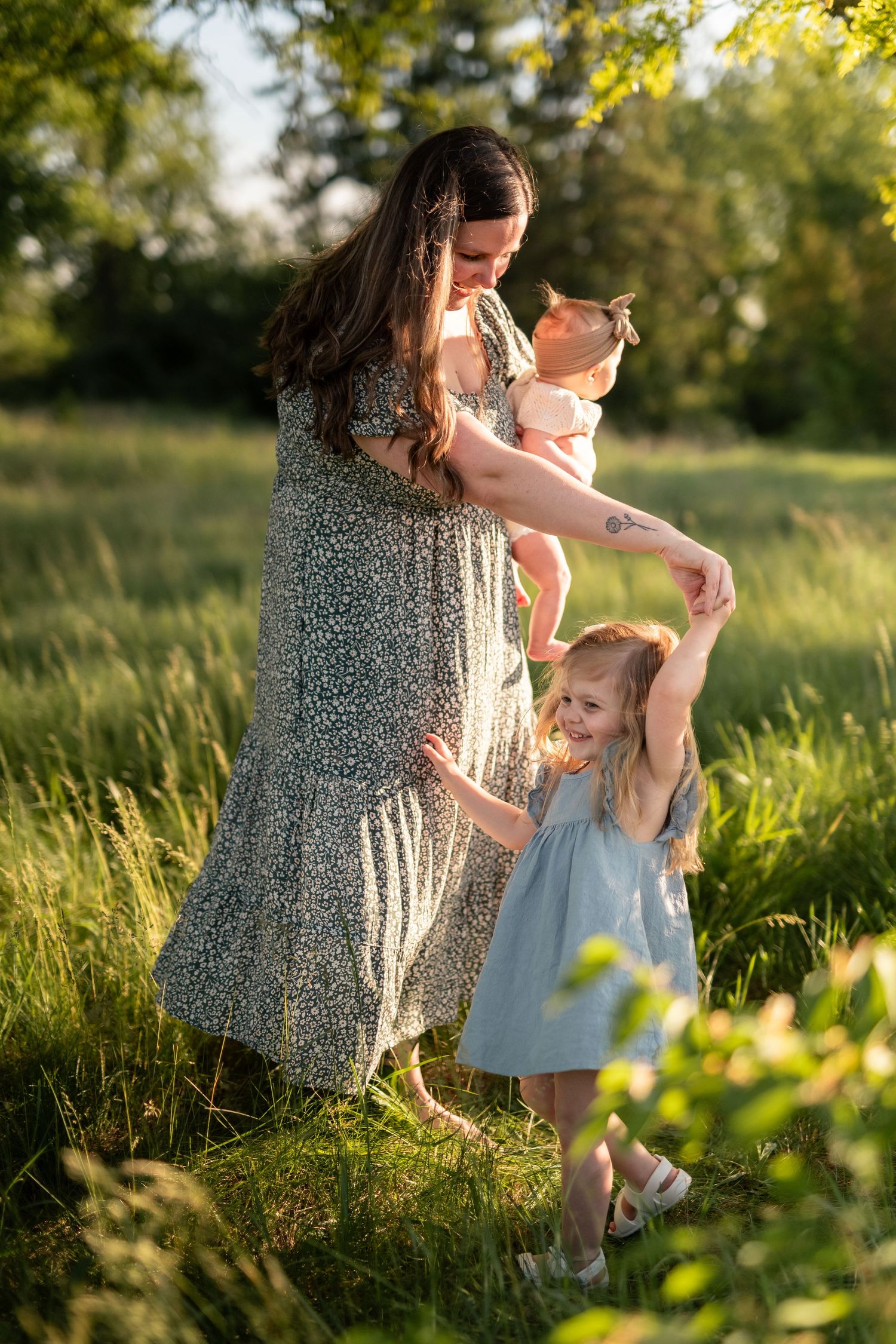 Mom cradles infant as smiling toddler girl dances outdoors in golden afternoon light.