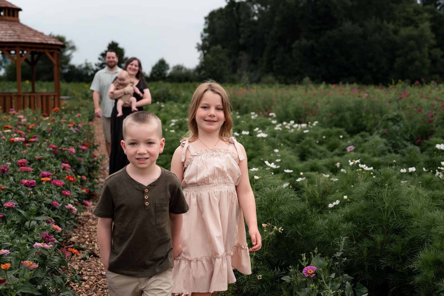 Two children smile in a flower garden path while parents with baby stand behind near a gazebo.
