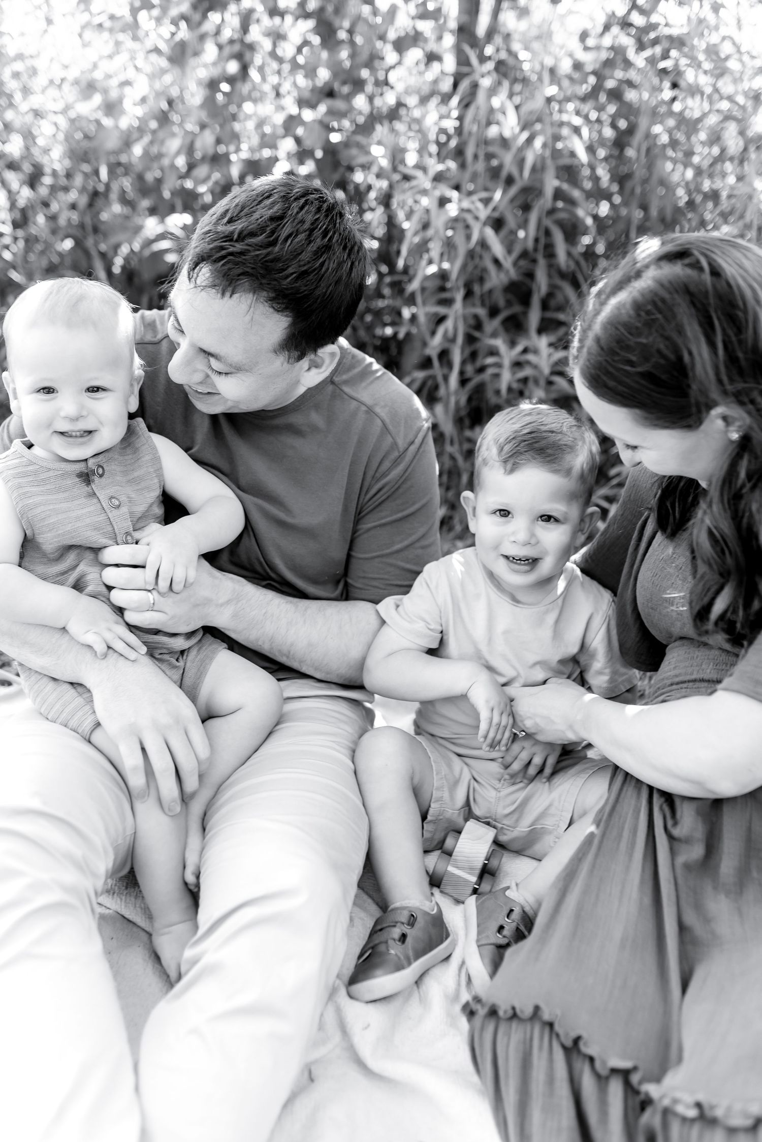 Black and white family portrait of parents with two young toddler boys sitting outdoors near tall grass.