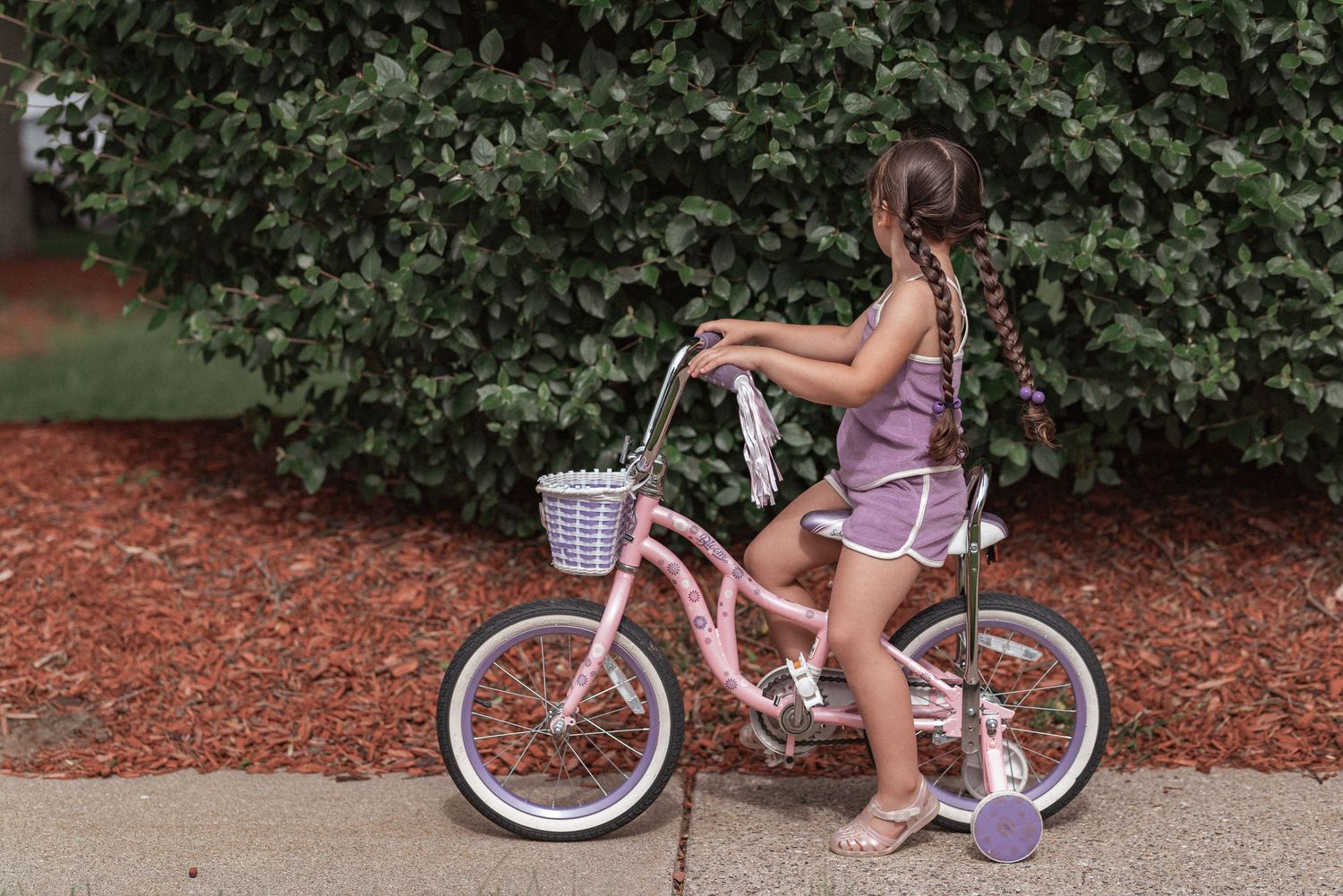 A young child learns to ride a pink balance bike along a tree-lined path with mulch ground cover.