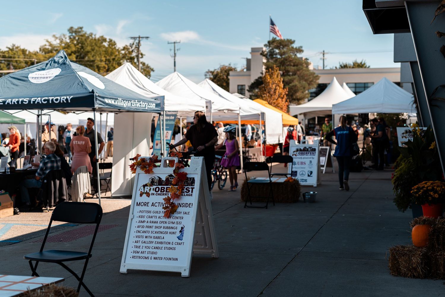 Outdoor arts festival with white vendor tents, shoppers browsing booths, and an American flag in the background on a sunny day.