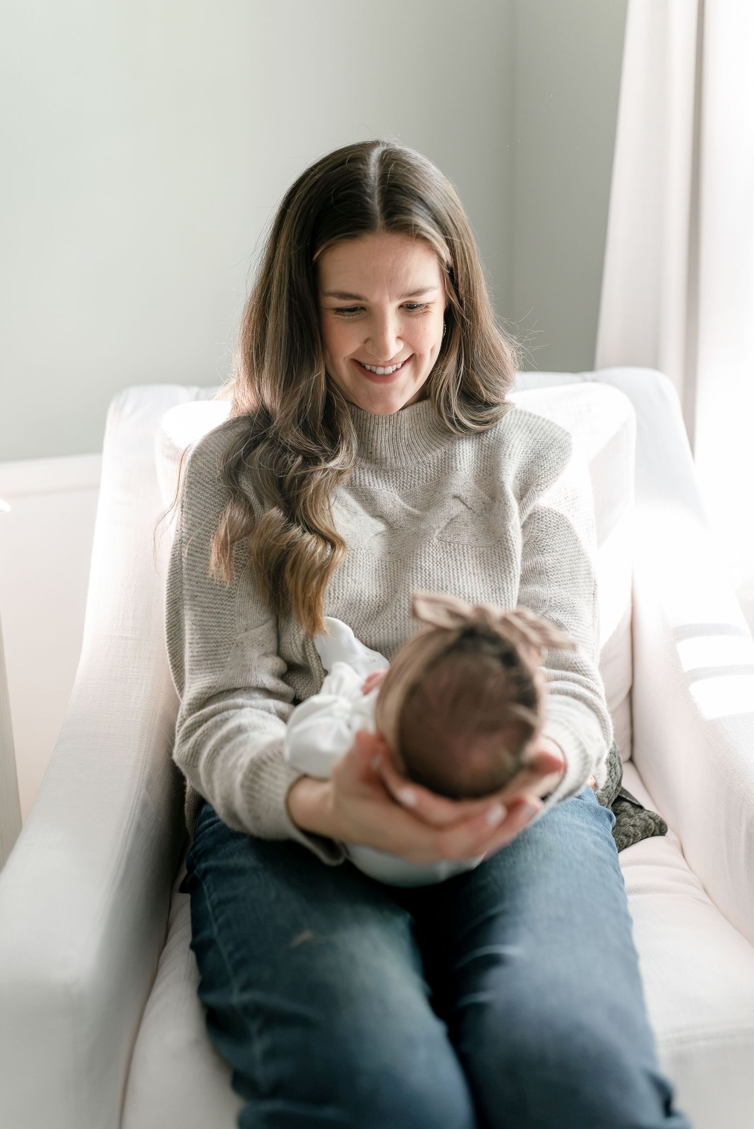 Smiling mother in gray sweater holding newborn baby while seated in white armchair.