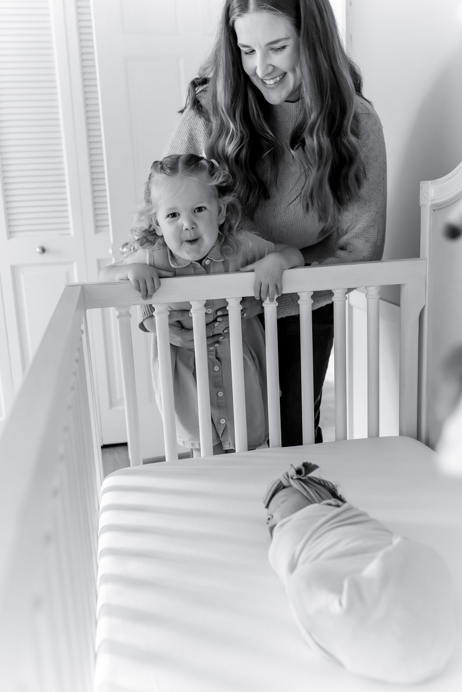 A smiling mother and toddler lean over a white crib to look at their swaddled newborn baby.