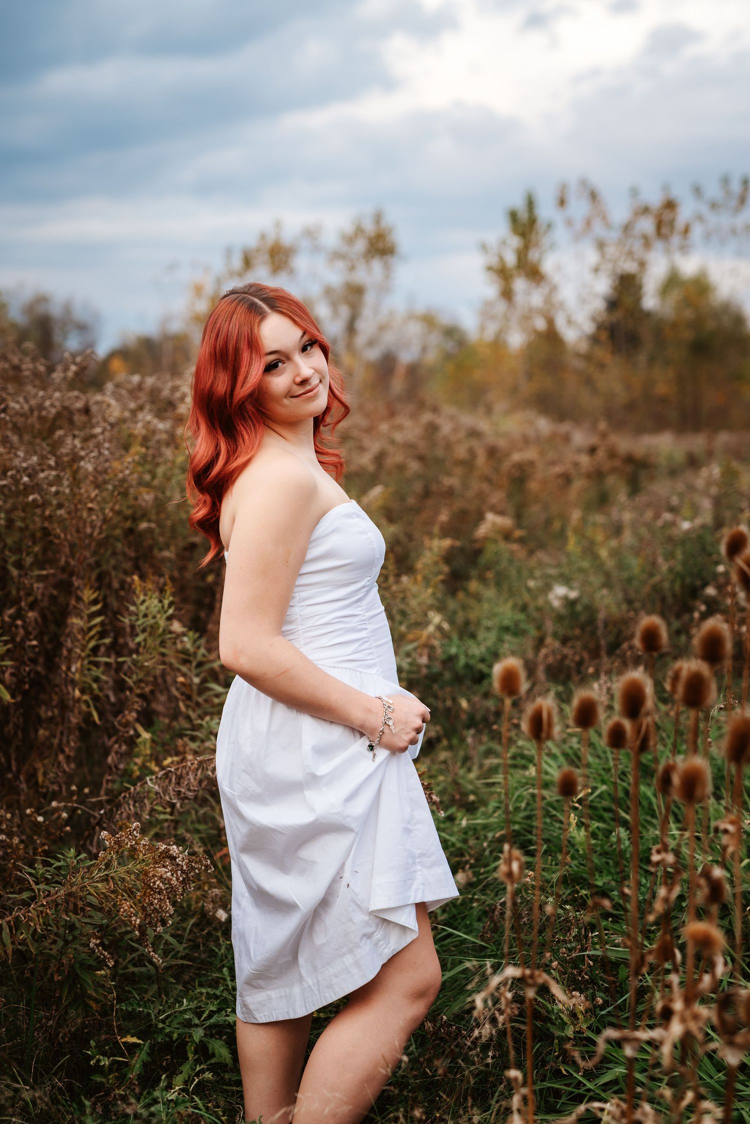 Young woman with red hair in white dress standing in autumn field with dried wildflowers under cloudy sky.