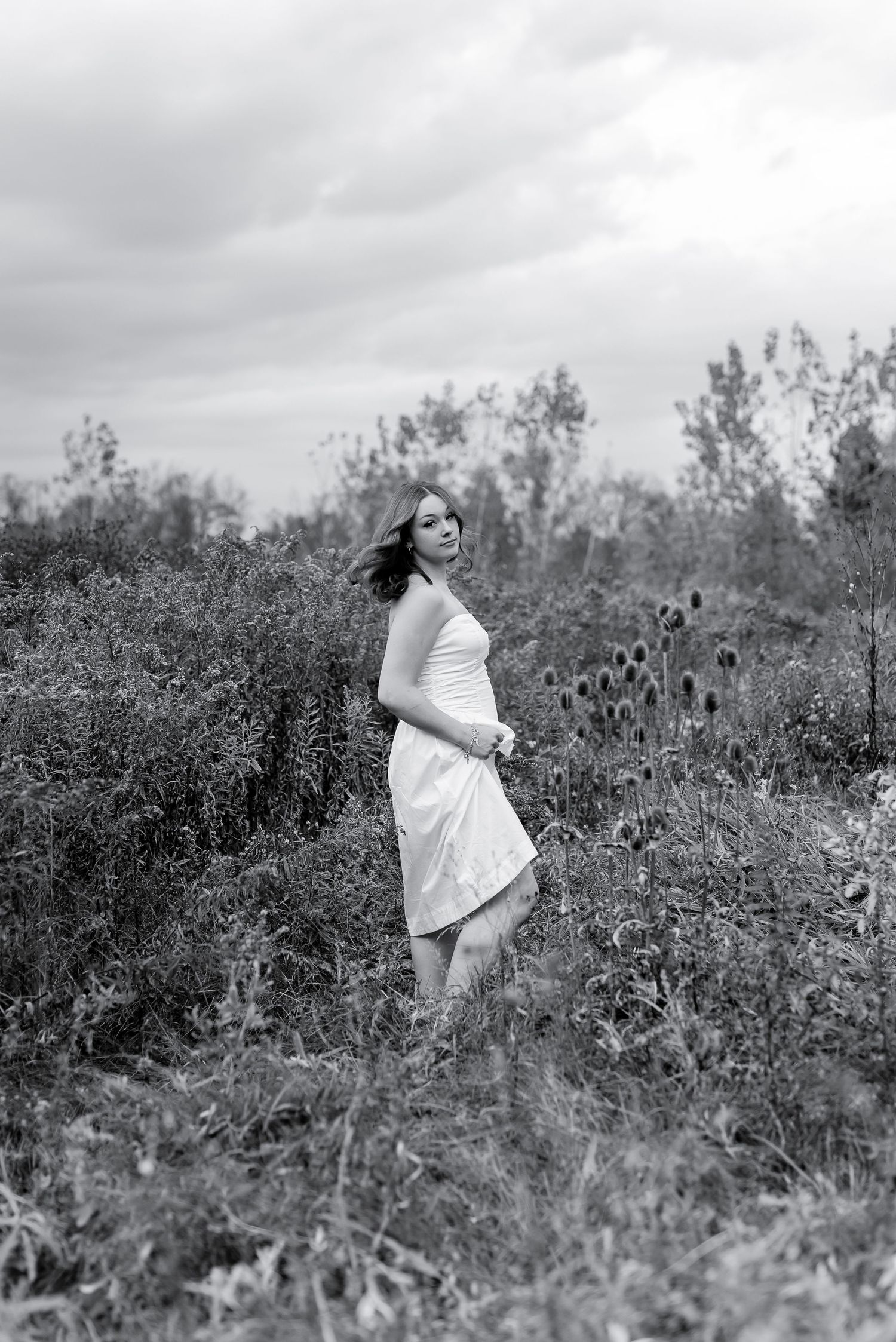 Black and white photo of young woman in white dress among wild plants and thistles in a field.