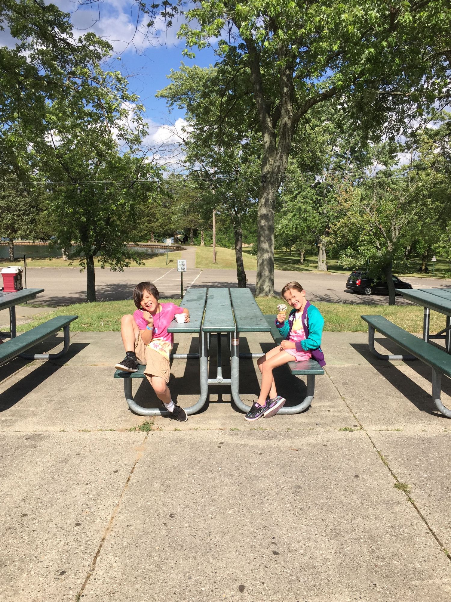 Two girls sitting at a green picnic table in a park surrounded by trees on a sunny day.