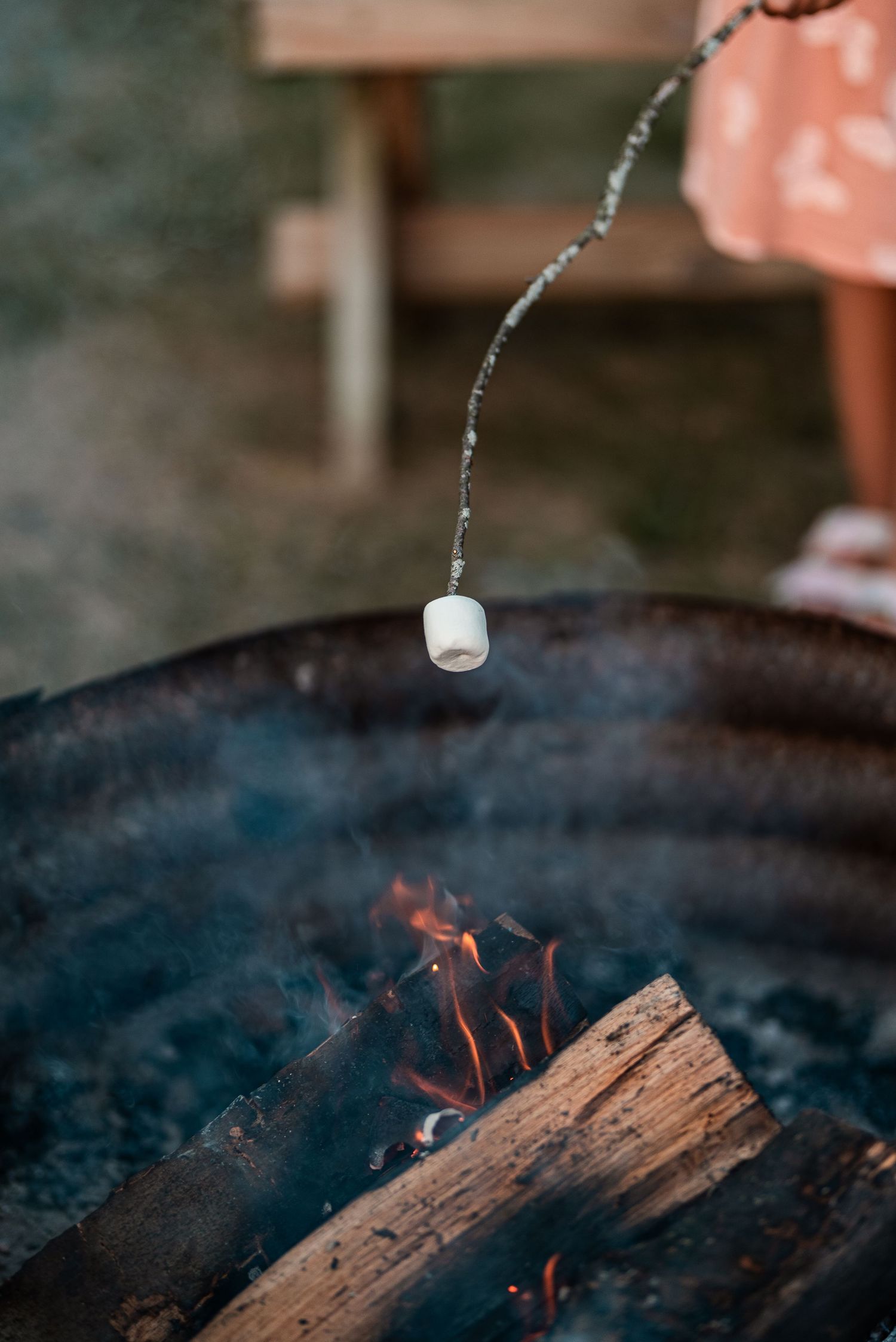 A marshmallow roasting on a stick over a campfire in a metal fire pit with glowing flames and smoke.