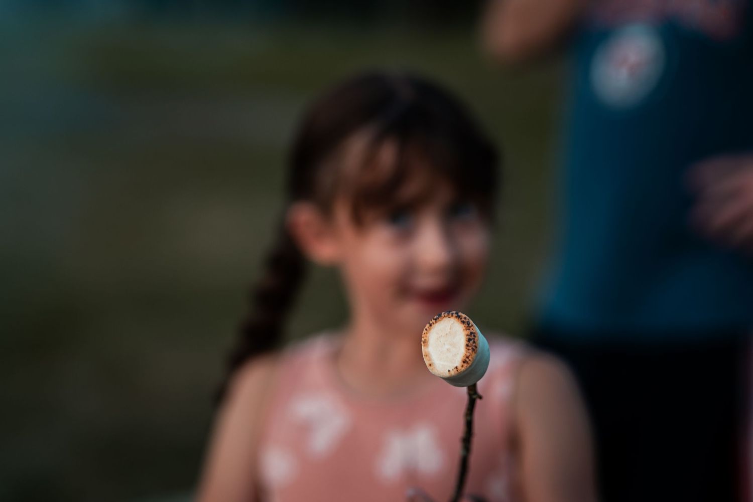 Close-up of toasted marshmallow on stick held by smiling girl with braided hair.