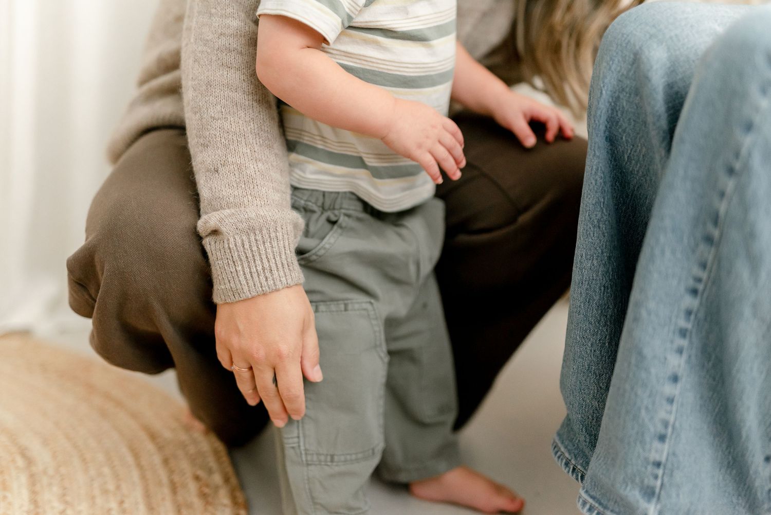 A toddler in gray pants stands between two adults in casual clothing in a bright, cozy indoor setting.