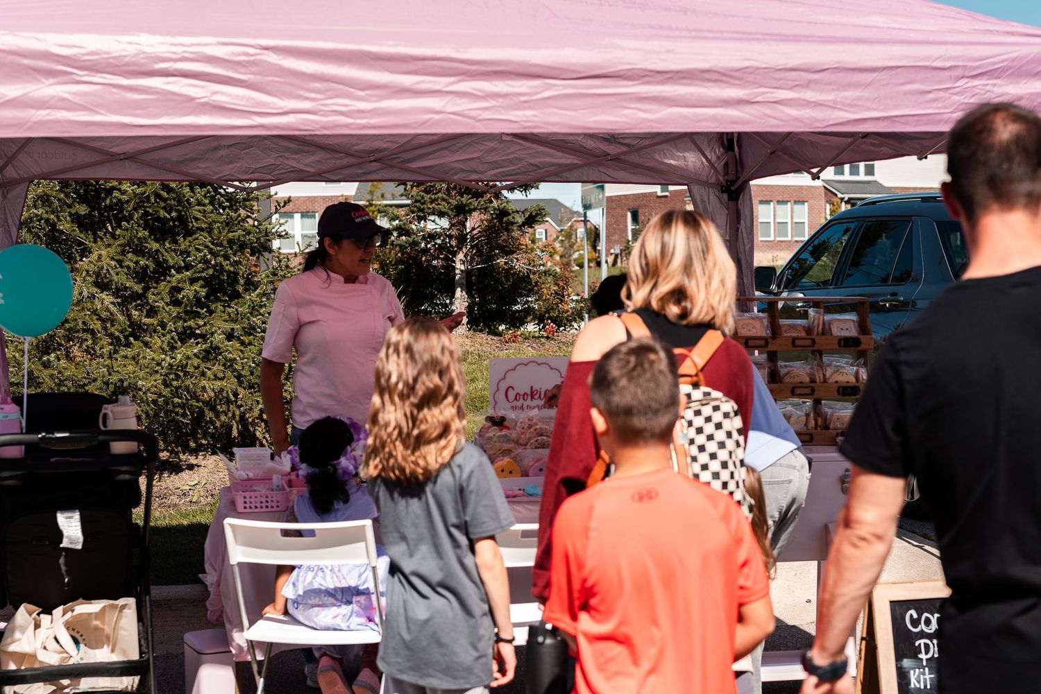 Families browsing a pink canopy vendor booth at a sunny outdoor community market event.