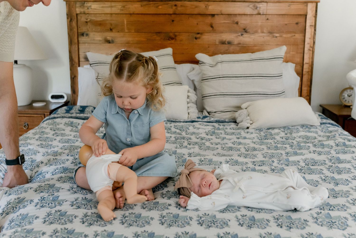 Toddler girl in blue dress sitting on bed next to sleeping newborn baby sibling.