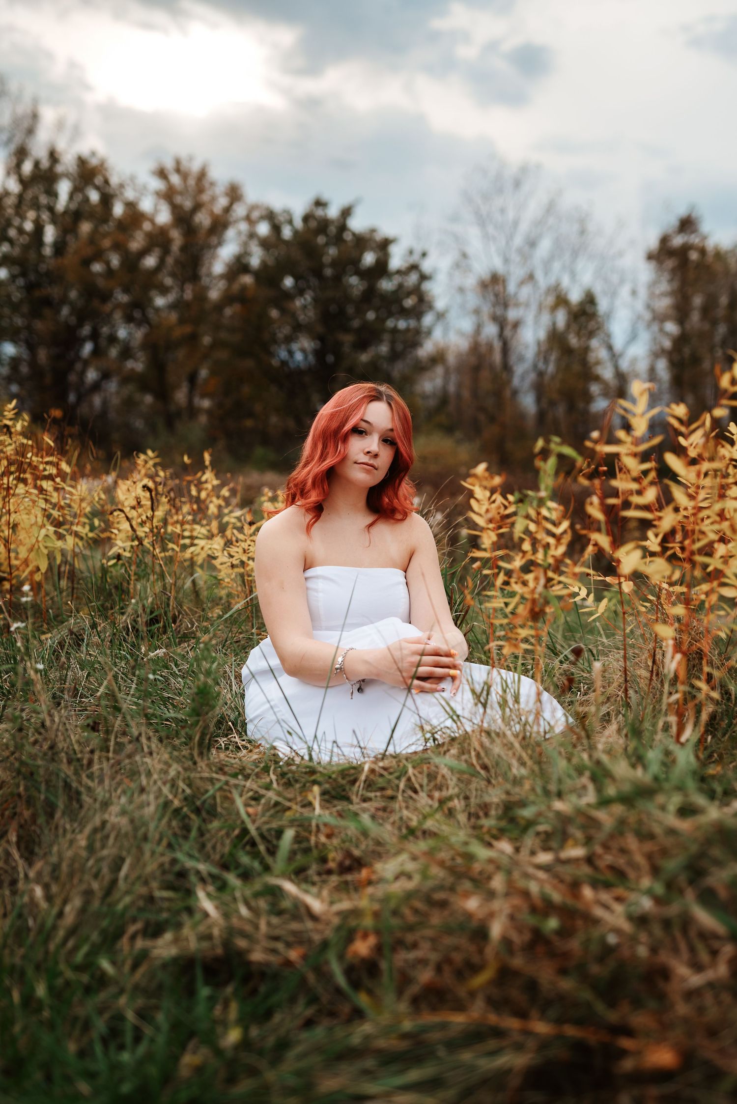 Red-haired woman in white strapless dress sitting peacefully in autumn field with golden foliage.