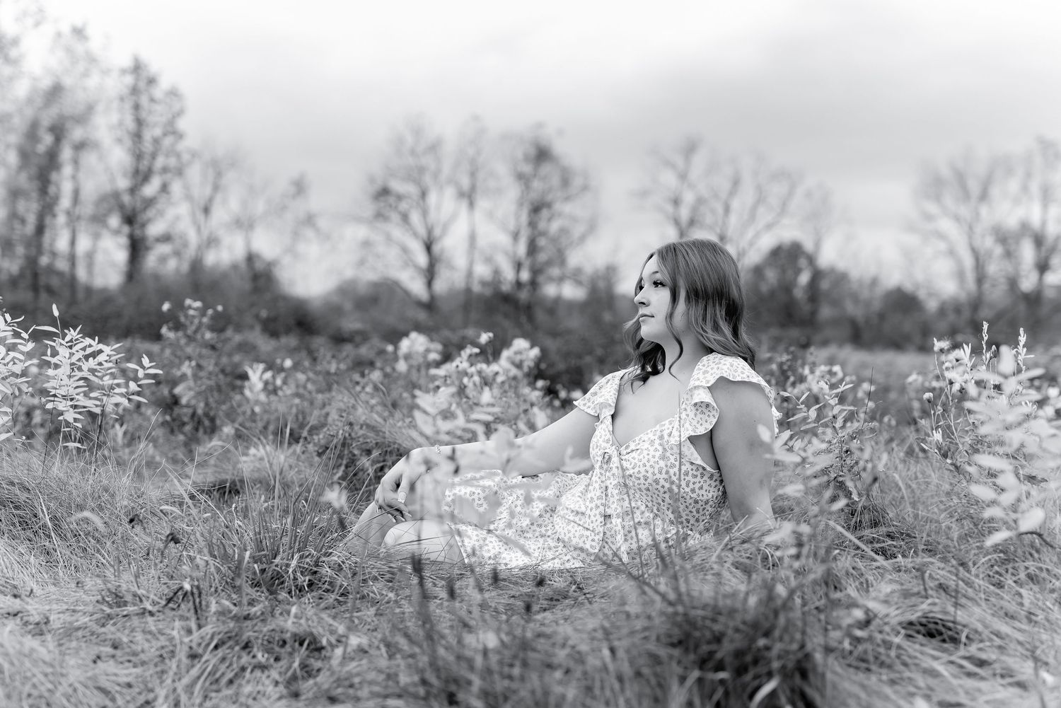 Black and white photo of young woman in floral dress sitting in a field gazing into distance.