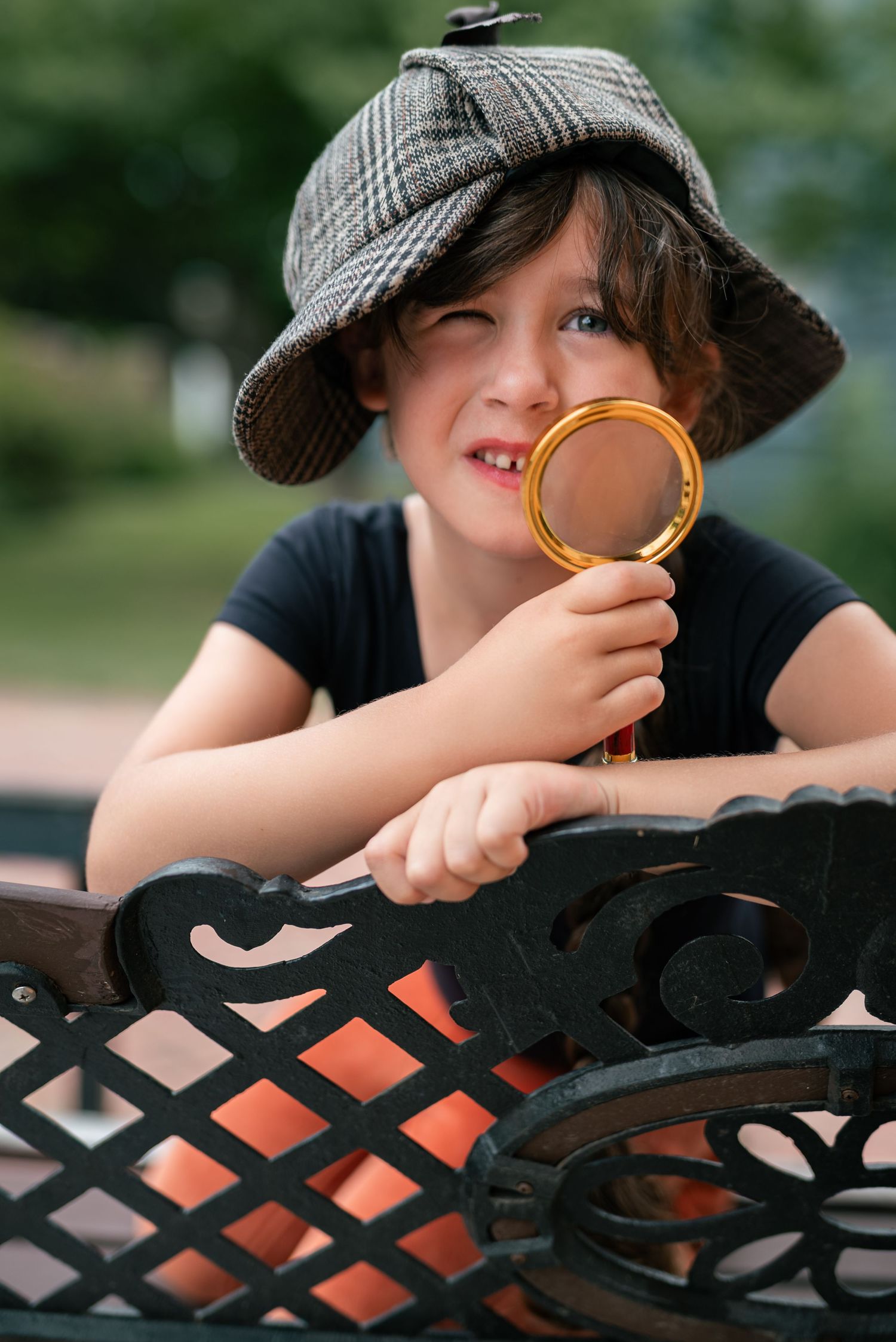 Child in deerstalker hat peers through golden magnifying glass over decorative iron park bench.