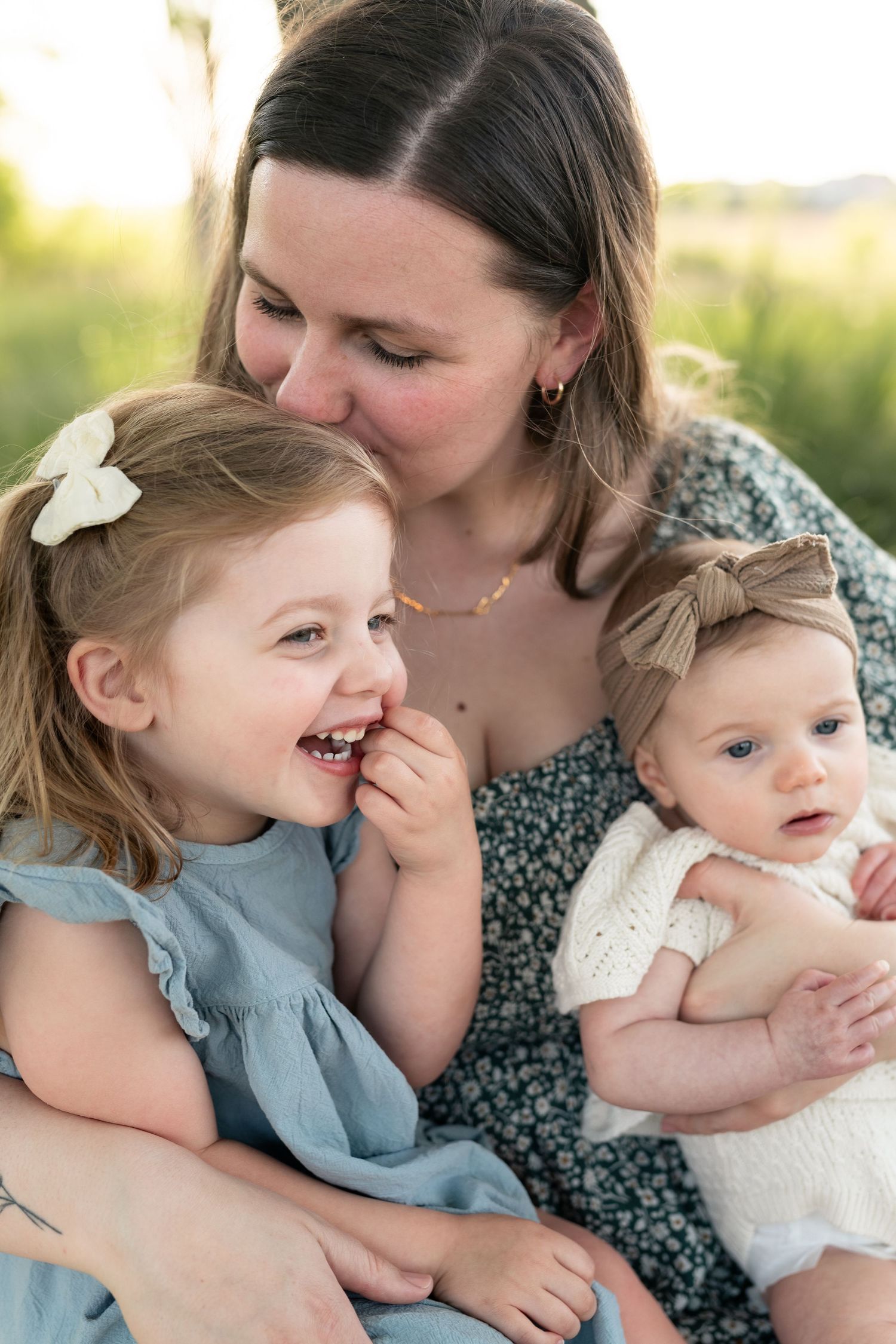 A mother smiles warmly at her two young daughters, a toddler girl and baby, sitting outdoors in soft golden light.
