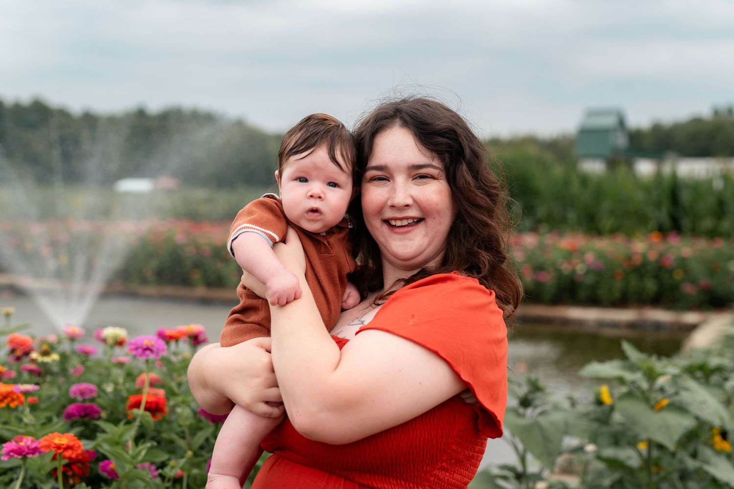 A smiling woman in a red top holds a baby near a garden fountain surrounded by colorful flowers outdoors.