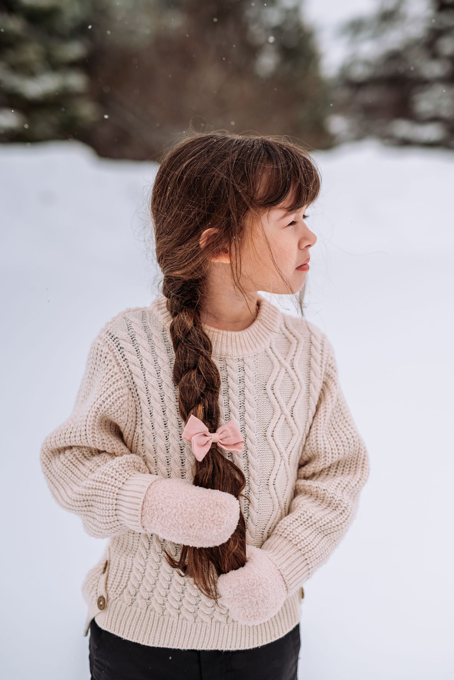A person wearing a cream cable knit sweater poses playfully in the snow with a pink bow in their braided hair.