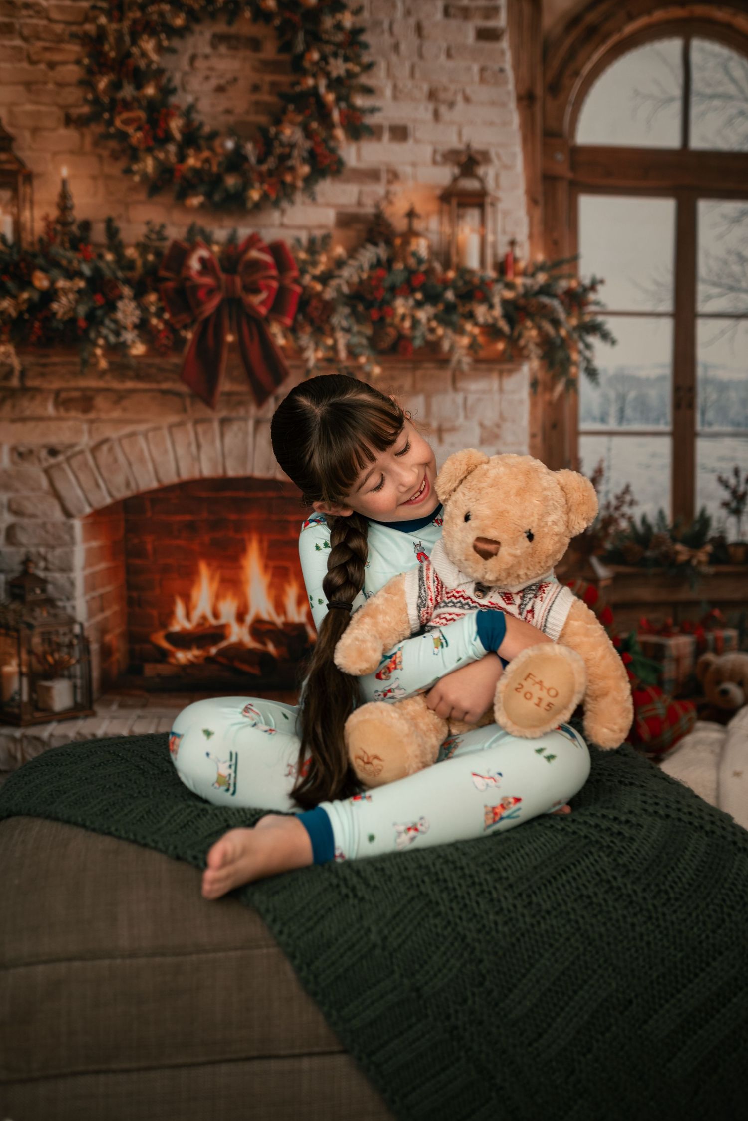 Girl in holiday pajamas hugs a teddy bear on a green knit blanket by a cozy brick fireplace with a garland and wreath.
