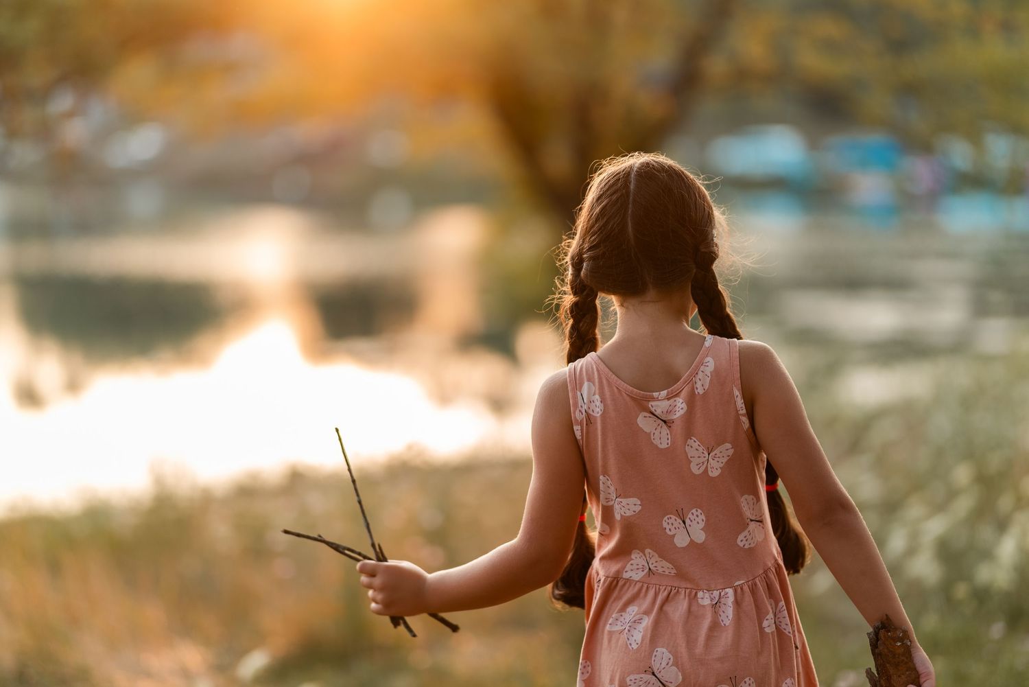 Young girl in pink floral dress holding a stick walks toward a sunlit lake at golden hour.