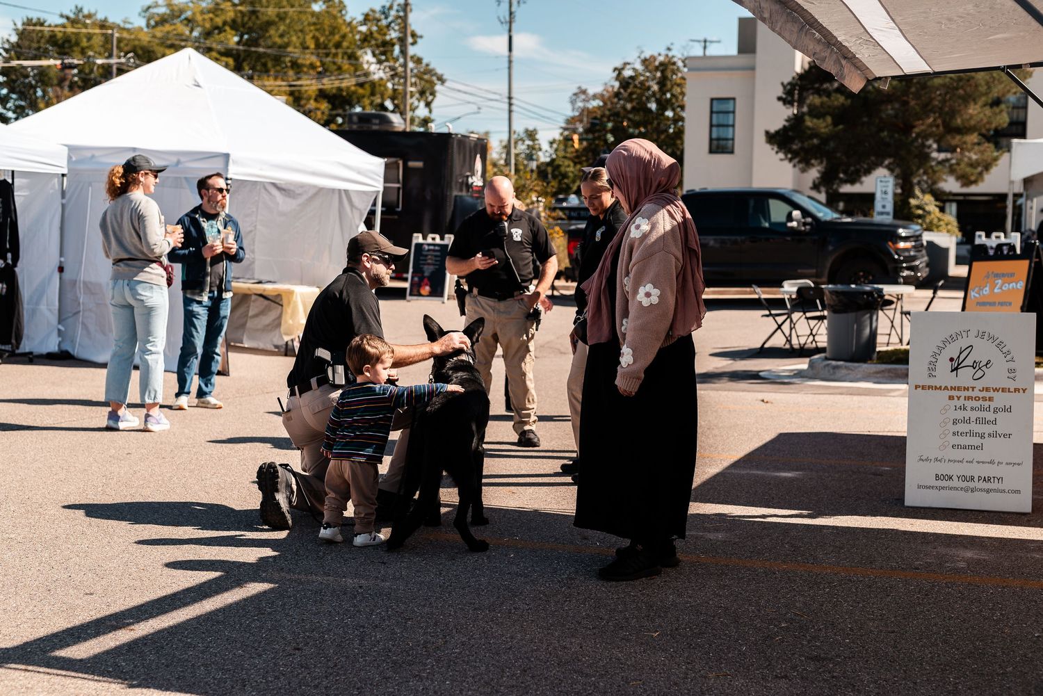 Outdoor market scene with people gathered around a vendor booth near white tents on a sunny day.