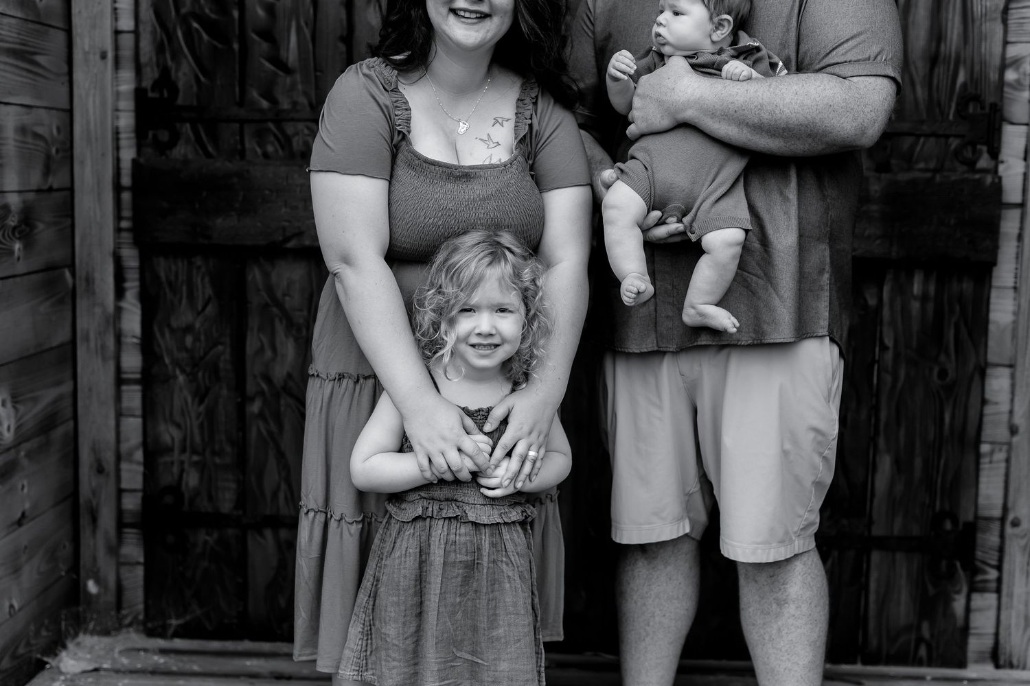 Black and white family portrait of parents with toddler girl and baby standing by a wooden door.