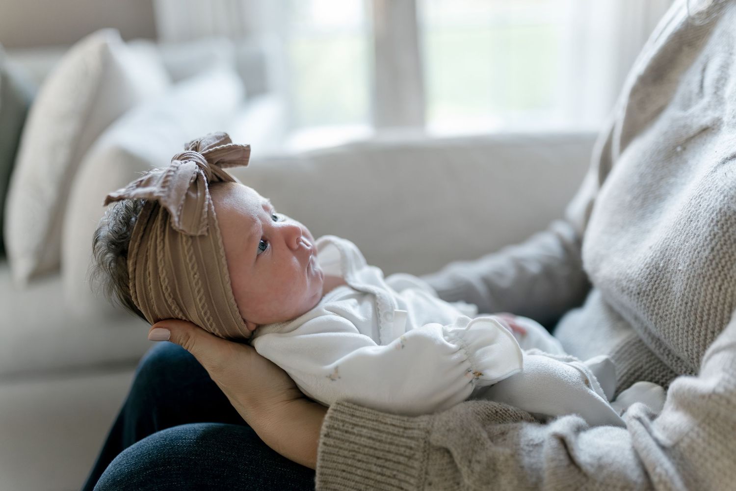 A newborn baby in a white onesie and bow headband rests peacefully in a parent's arms on a couch.