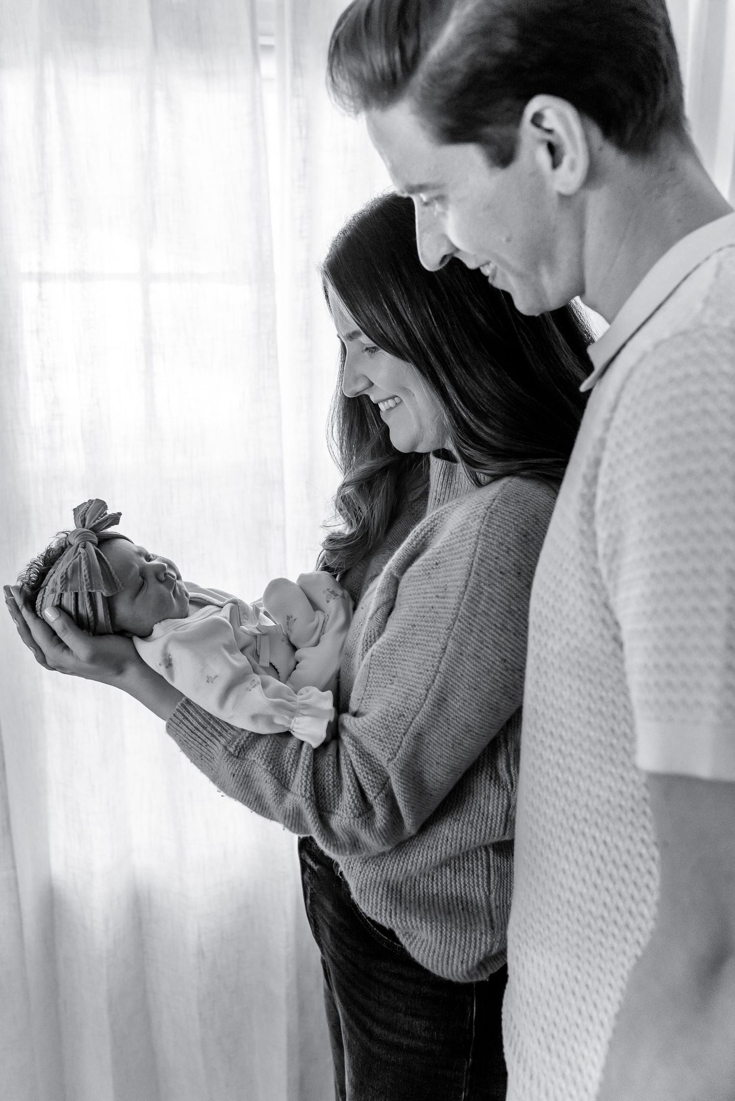 Black and white photo of new parents lovingly gazing at their newborn baby by a sheer curtained window.