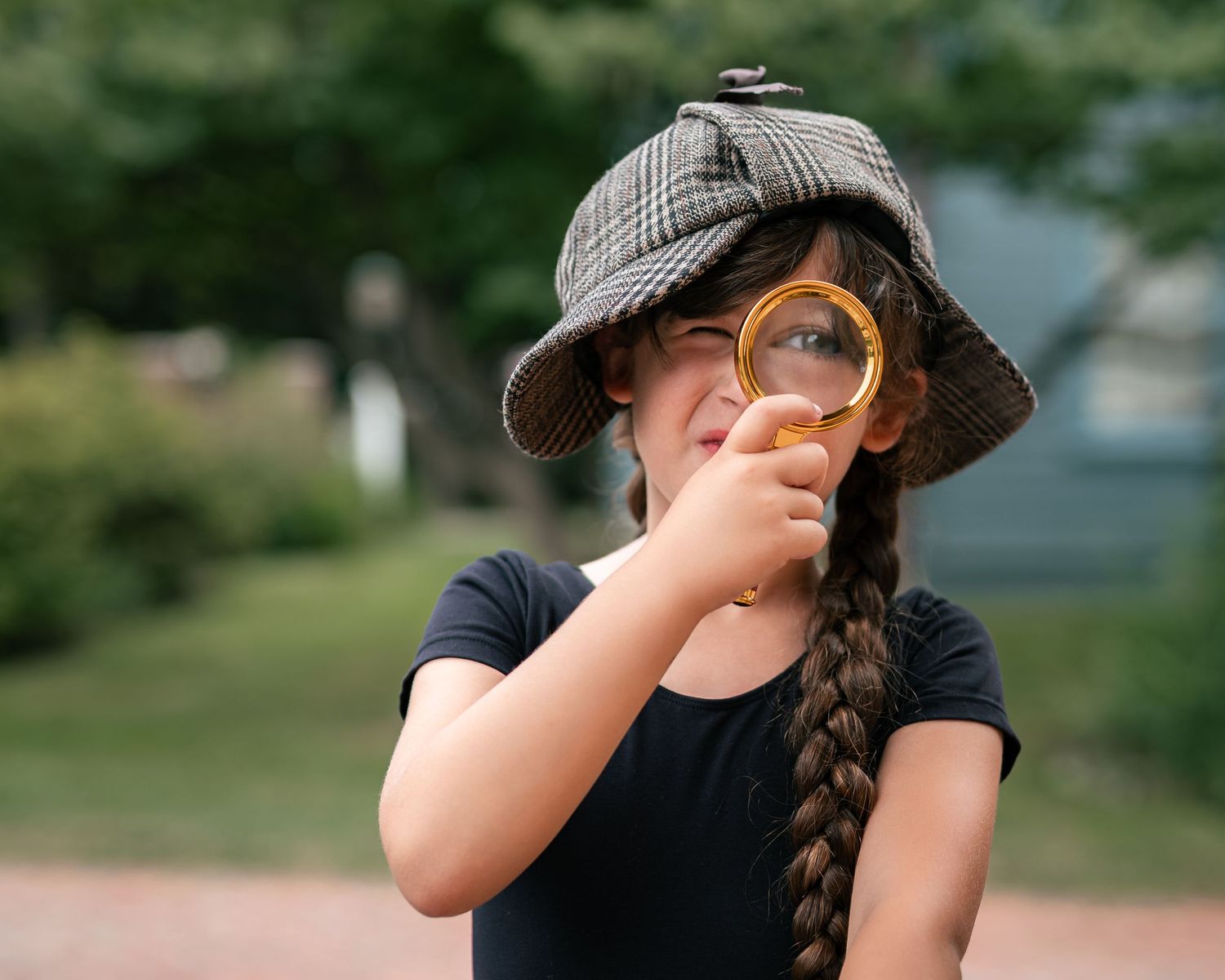 Young girl in plaid detective hat holds magnifying glass to her eye outdoors, playing inspector.