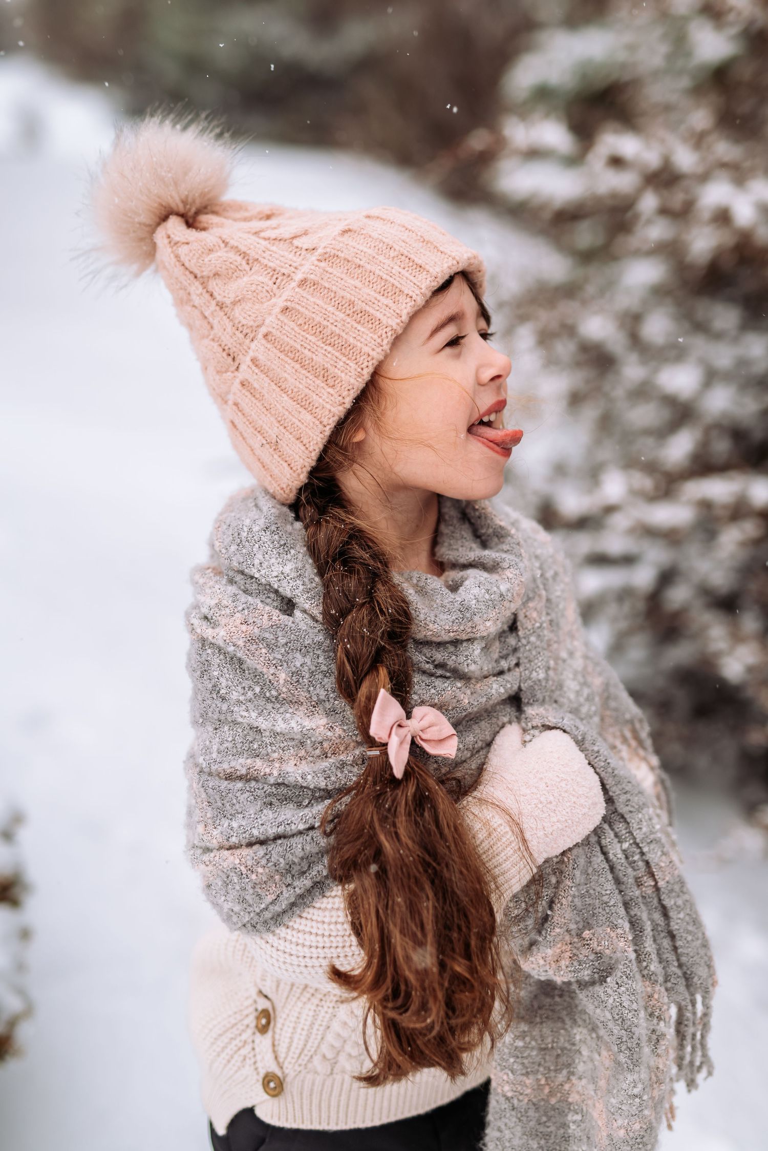 A winter fashion look featuring a pink knit beanie, gray scarf, and long brown hair in a snowy outdoor setting.