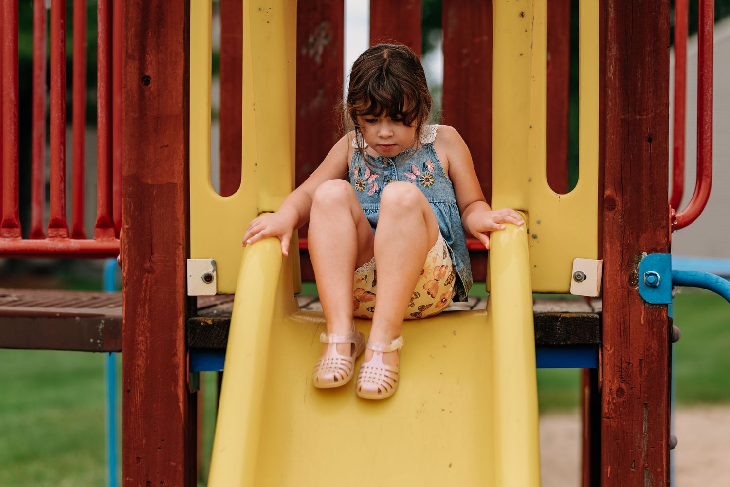 Child playing on a yellow playground slide with red supports during a summer day at the park.