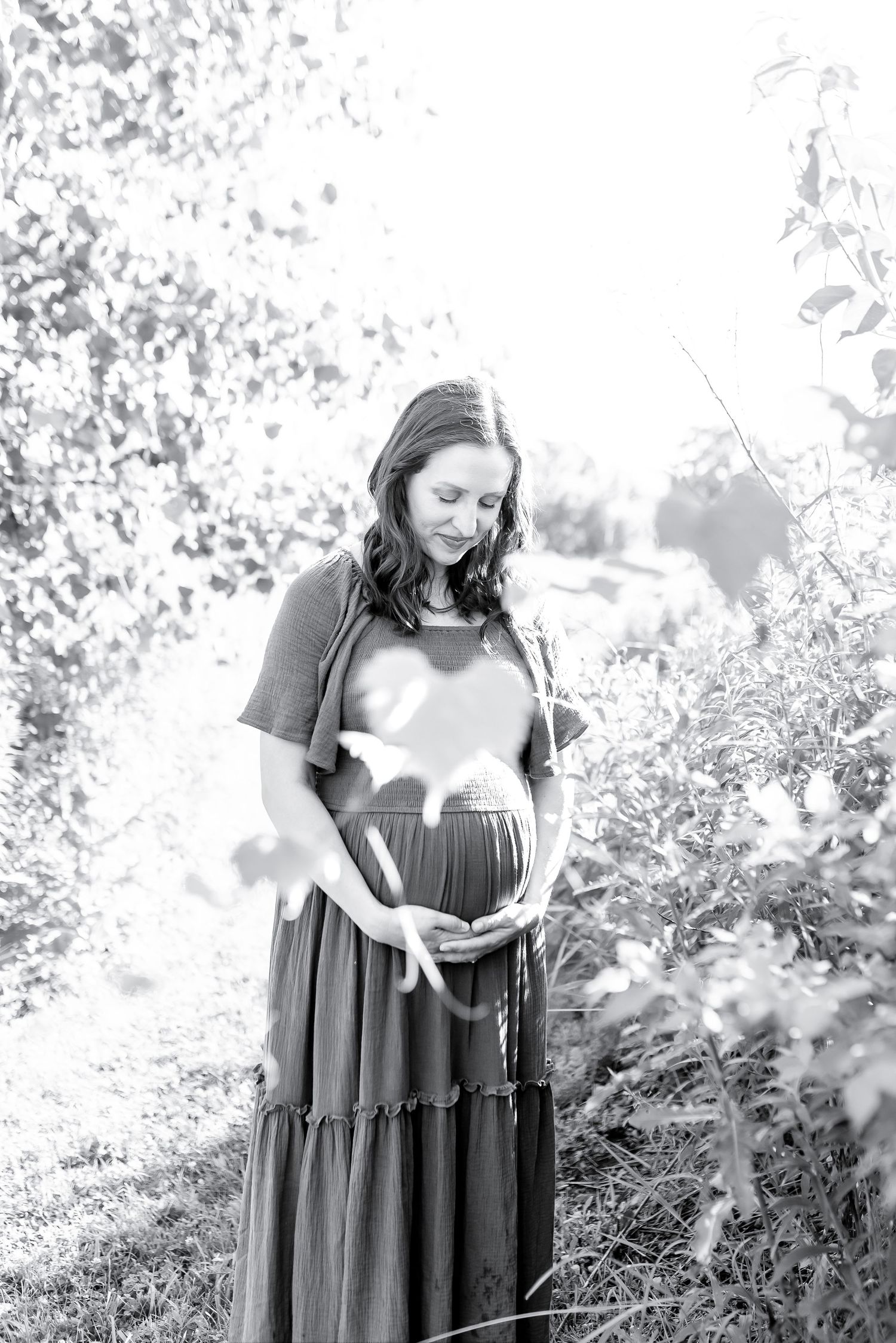 Black and white outdoor maternity photo session showing mother in flowing dress surrounded by foliage.