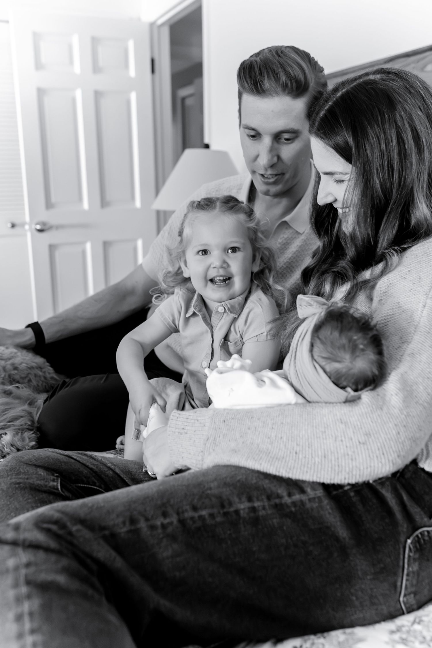 Black and white family portrait of parents with toddler and newborn baby sitting on bed.