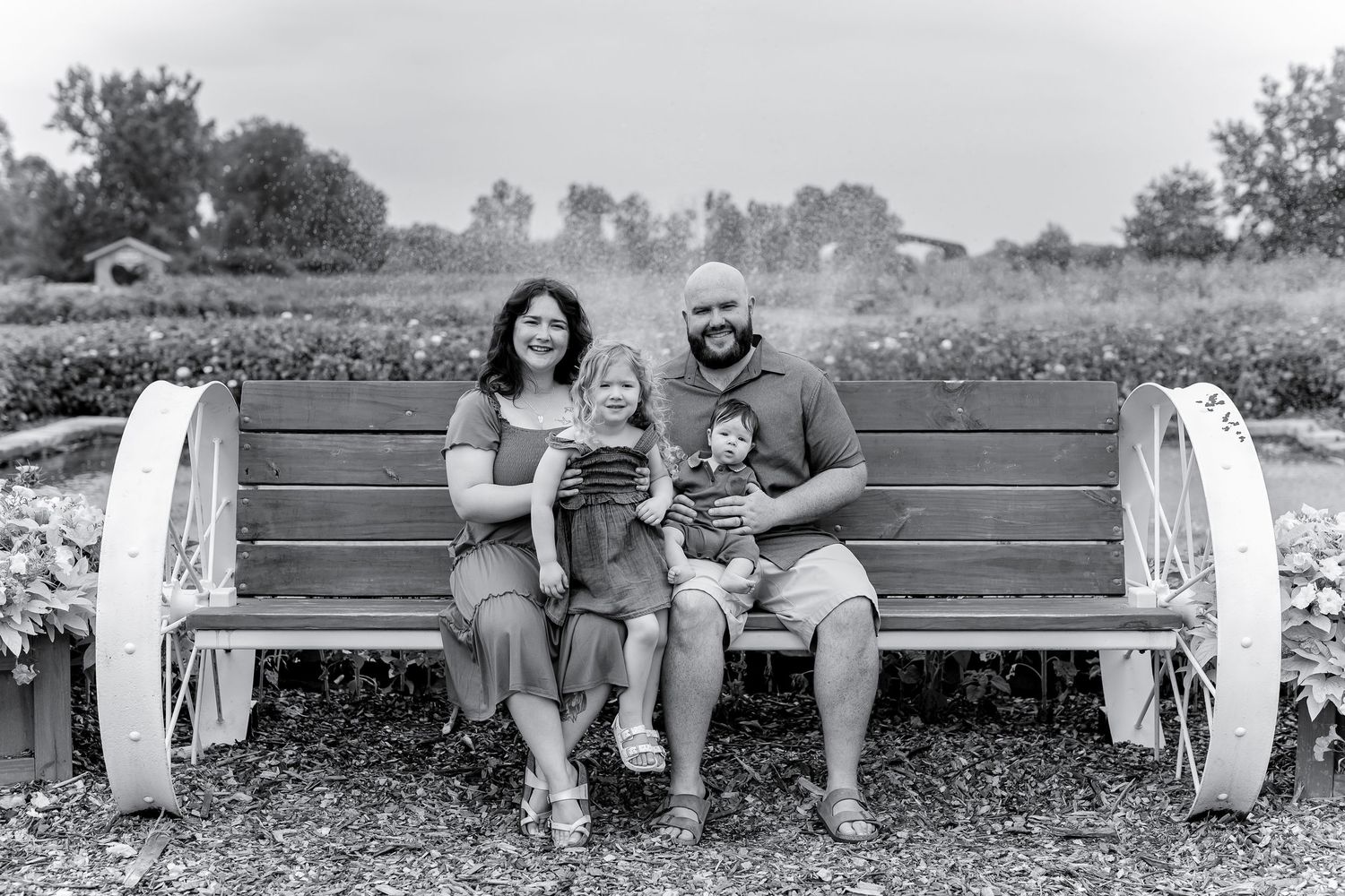 Black and white family portrait of four sitting on decorative wagon wheel bench.