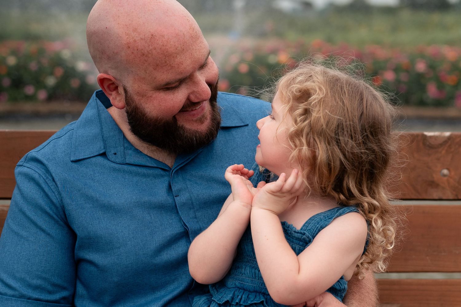 A bald bearded father in blue shirt smiles at his curly-haired toddler daughter on a wooden bench outdoors.