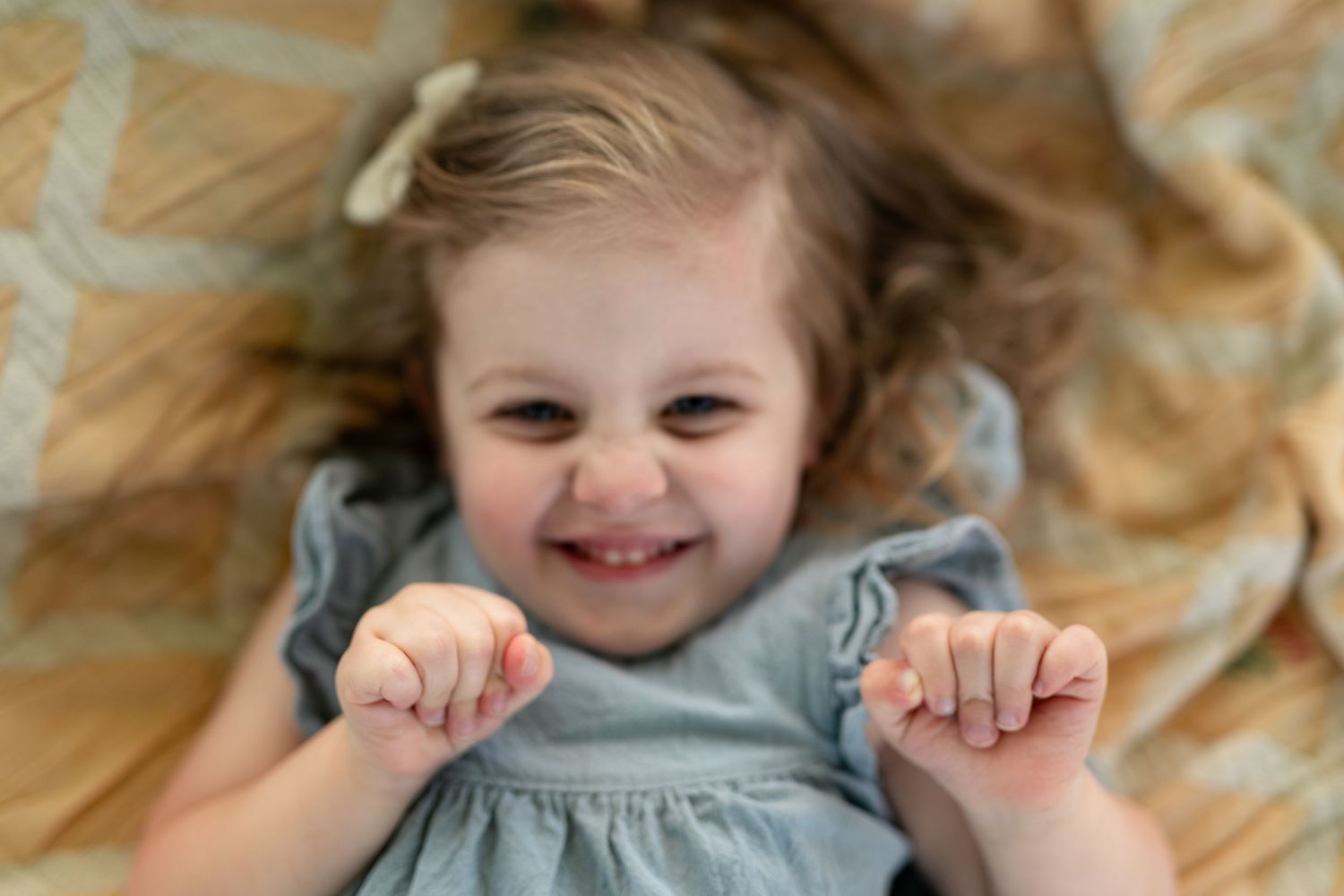 Smiling toddler girl in gray dress lying on blanket, hands raised, with a white bow in her hair.