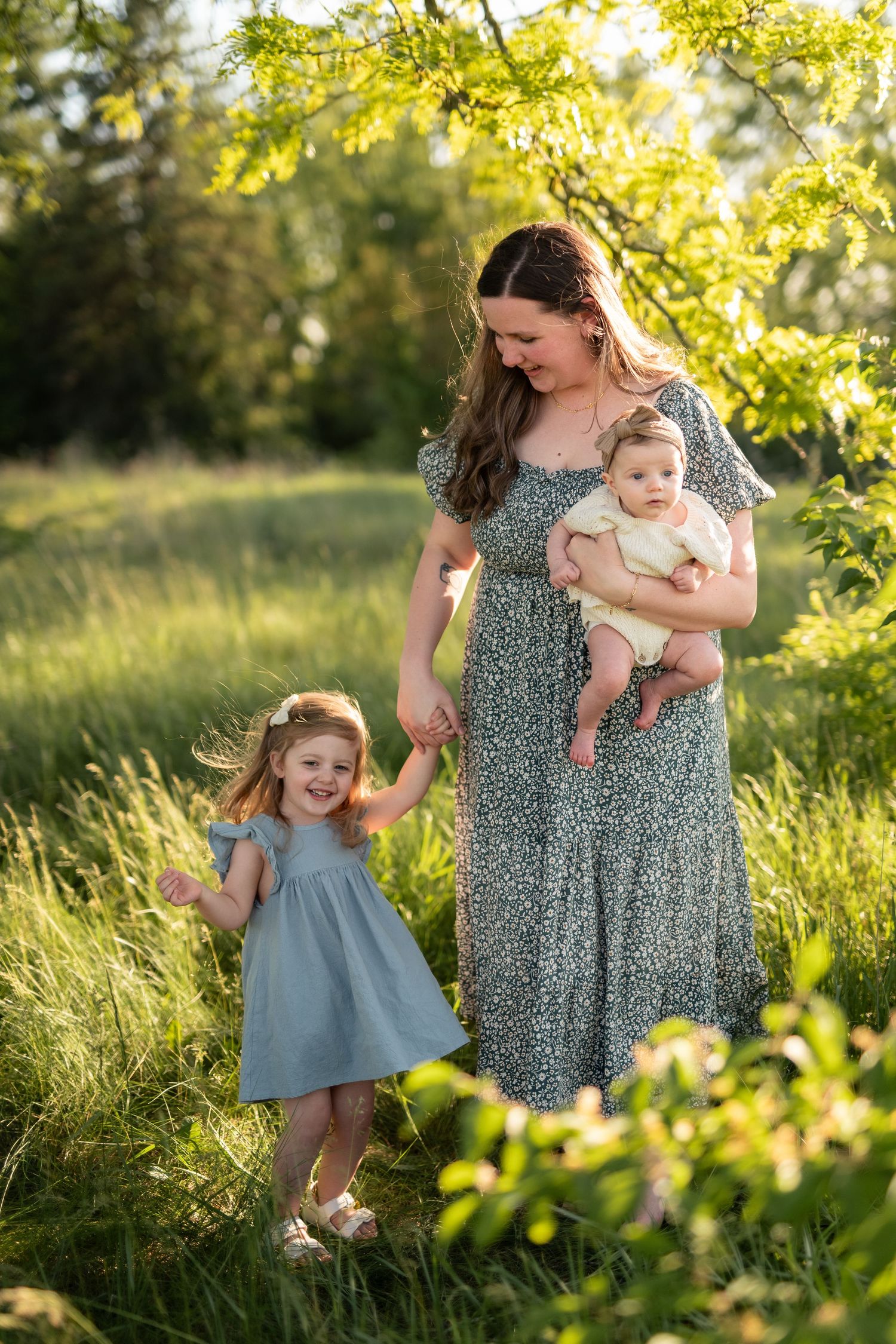 A mother in a floral dress holds her baby while her young daughter in a blue dress smiles outdoors in a sunny green field.