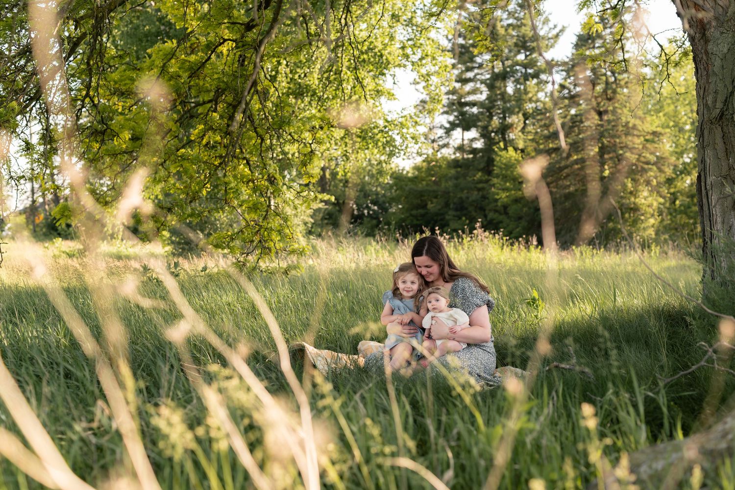 A mother sits with two young children in a lush green meadow surrounded by tall grass and trees on a sunny day.