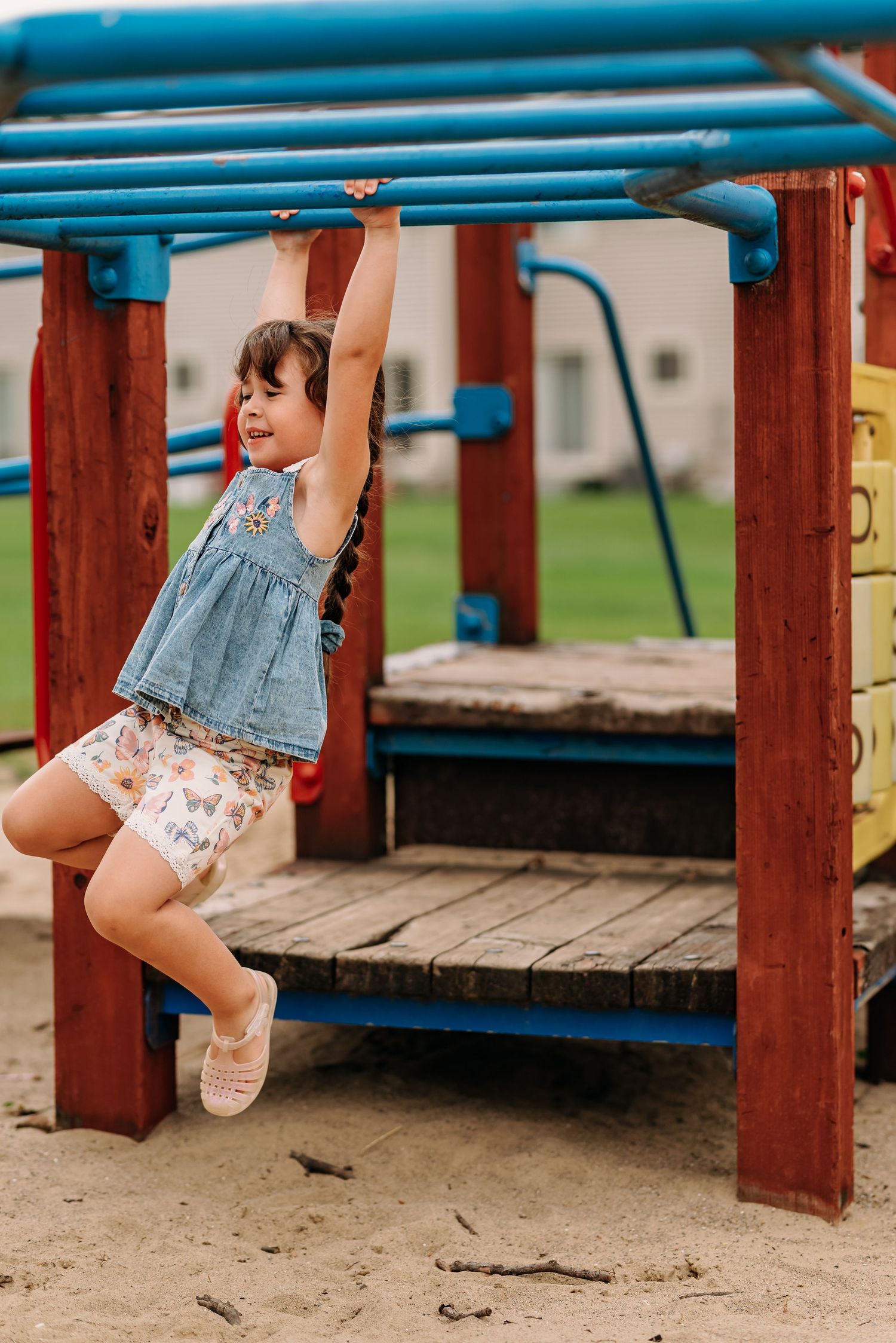 A joyful child swinging on blue monkey bars at a playground wearing denim and floral clothing.