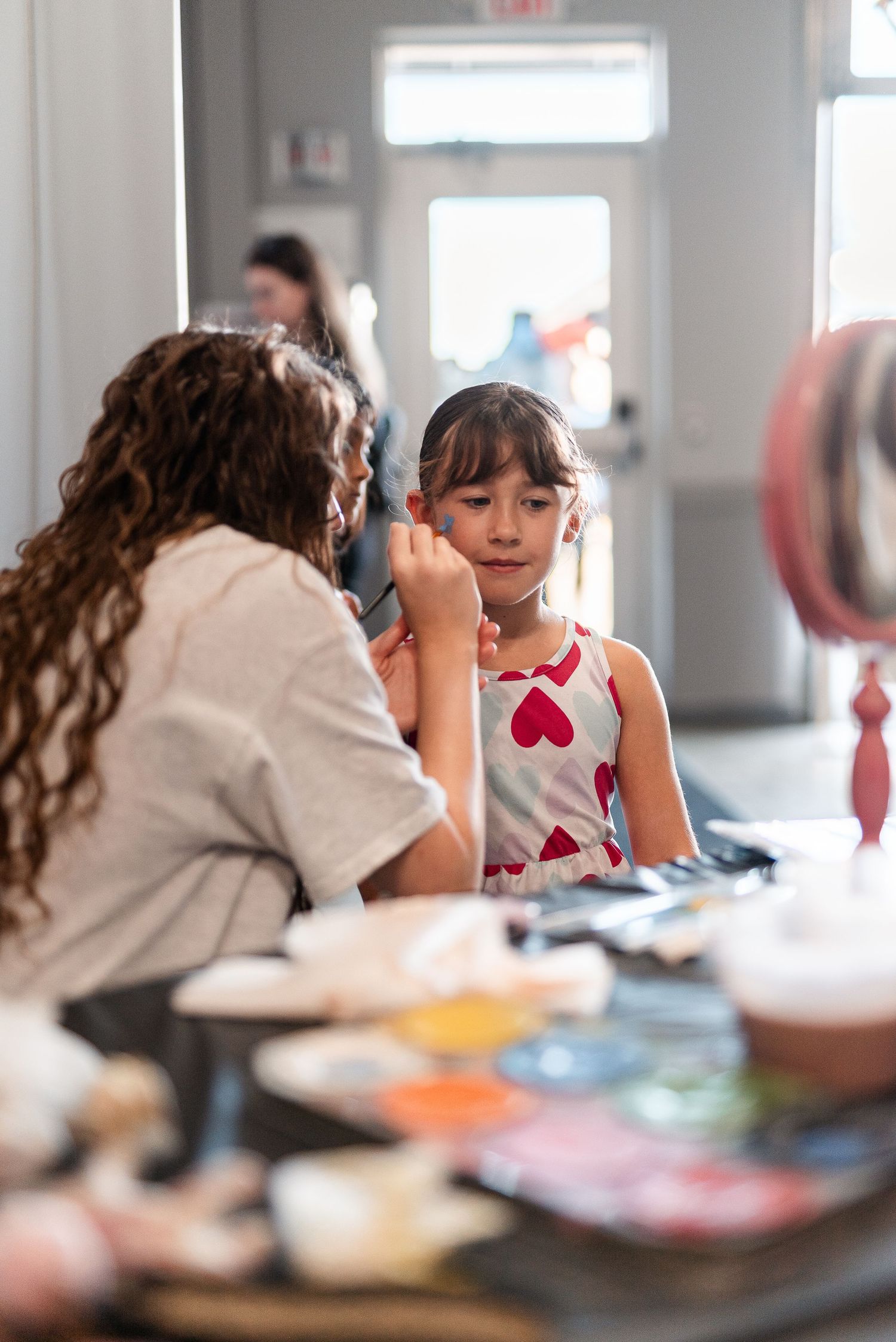 A woman applies face paint to a young girl at a colorful face painting station indoors.