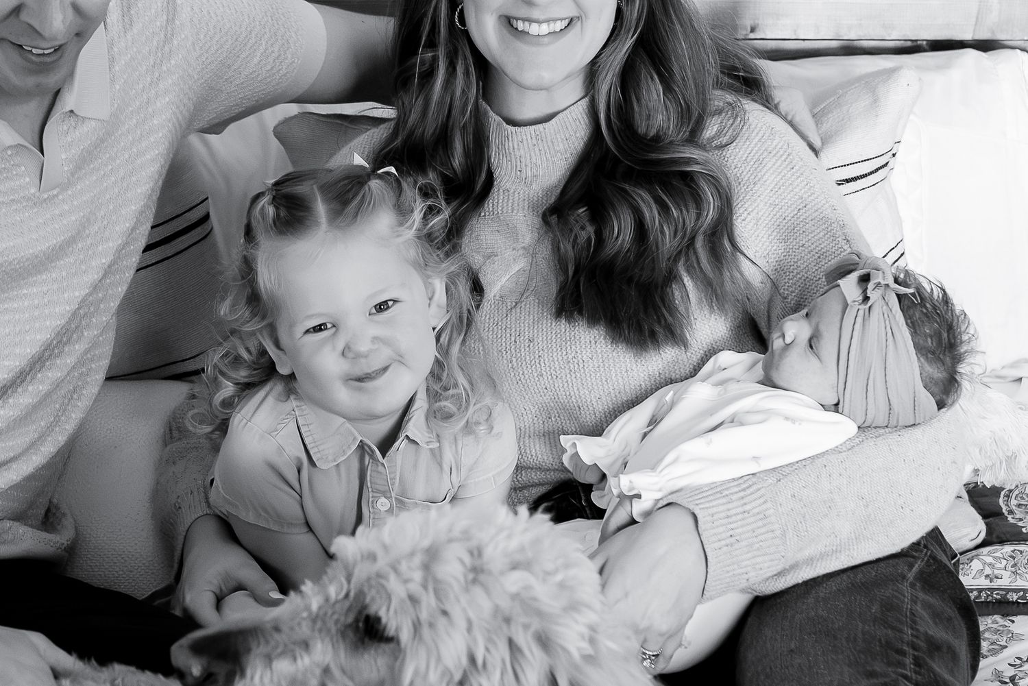 Black and white family photo with mother holding newborn, young daughter, dog, and father on a cozy bed.