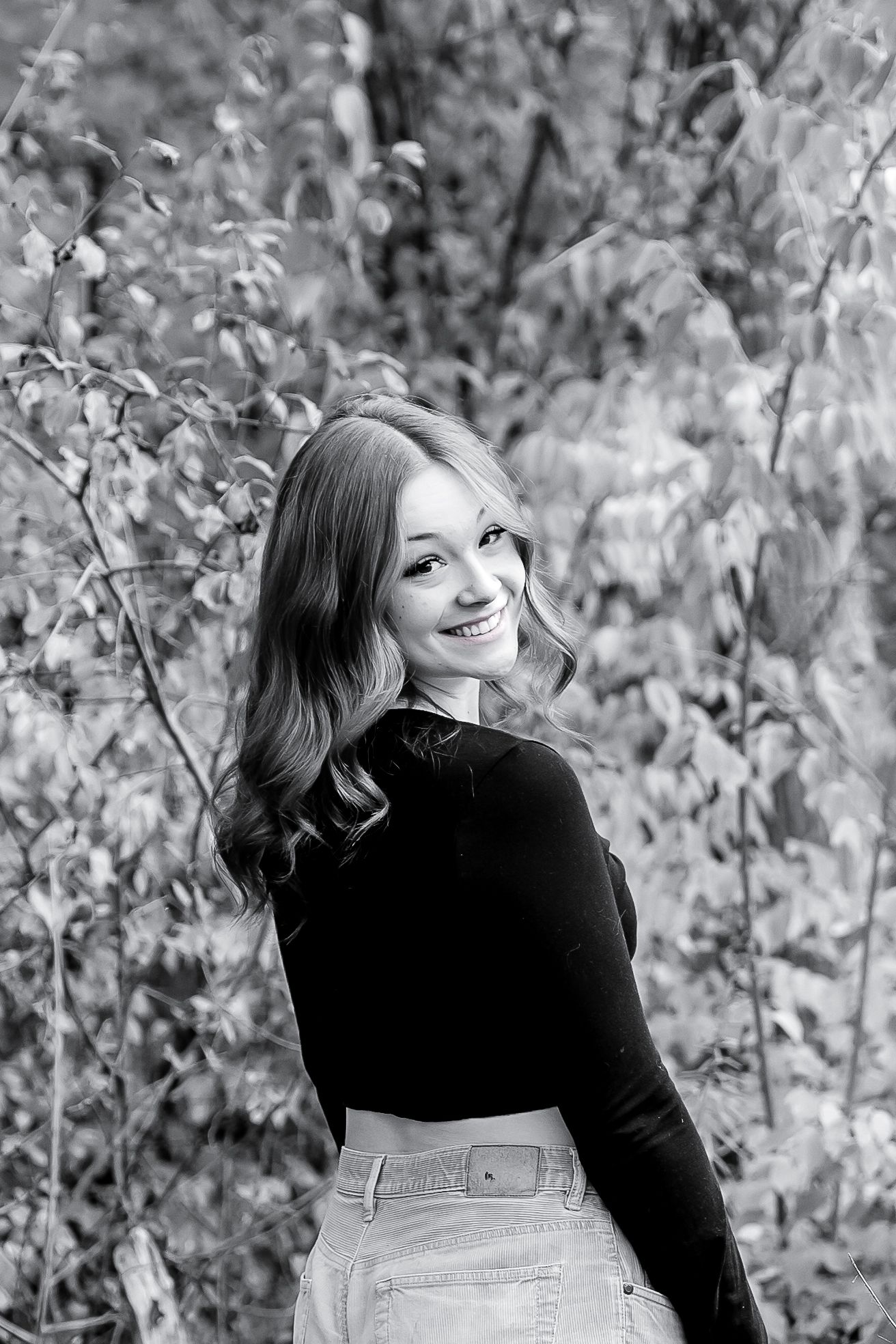 Black and white portrait of a smiling young woman in a dark top standing outdoors among leafy trees.