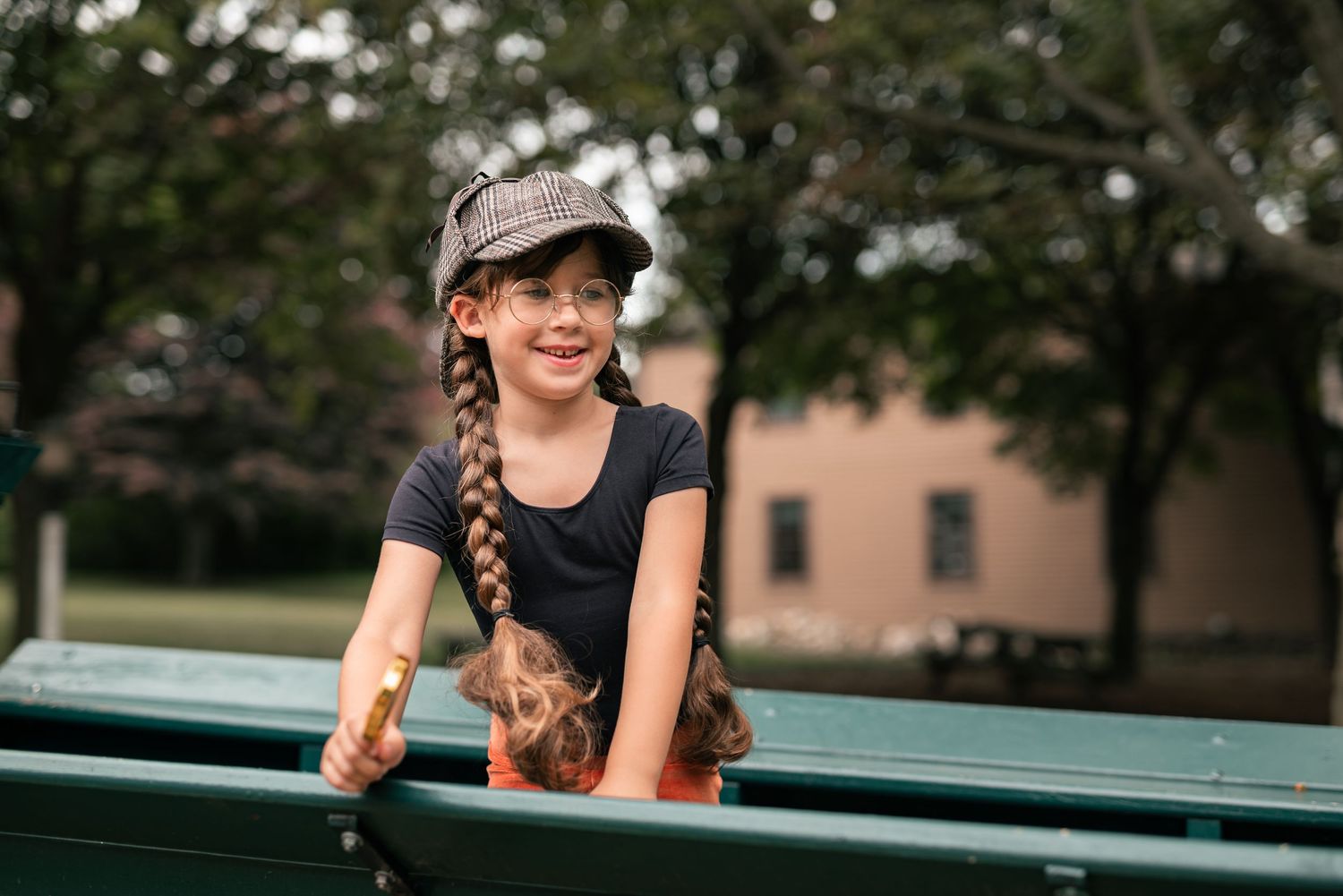 A smiling young girl with braids and a plaid cap sits on a green park bench outdoors.