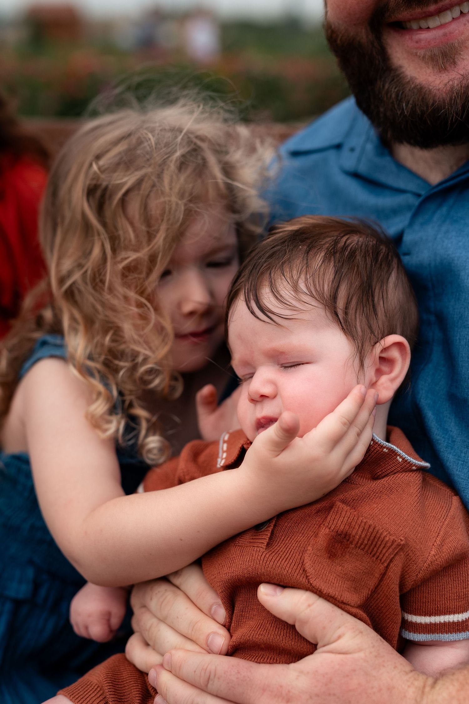 A curly-haired toddler girl gently touches a sleeping newborn baby held by a bearded man in blue.